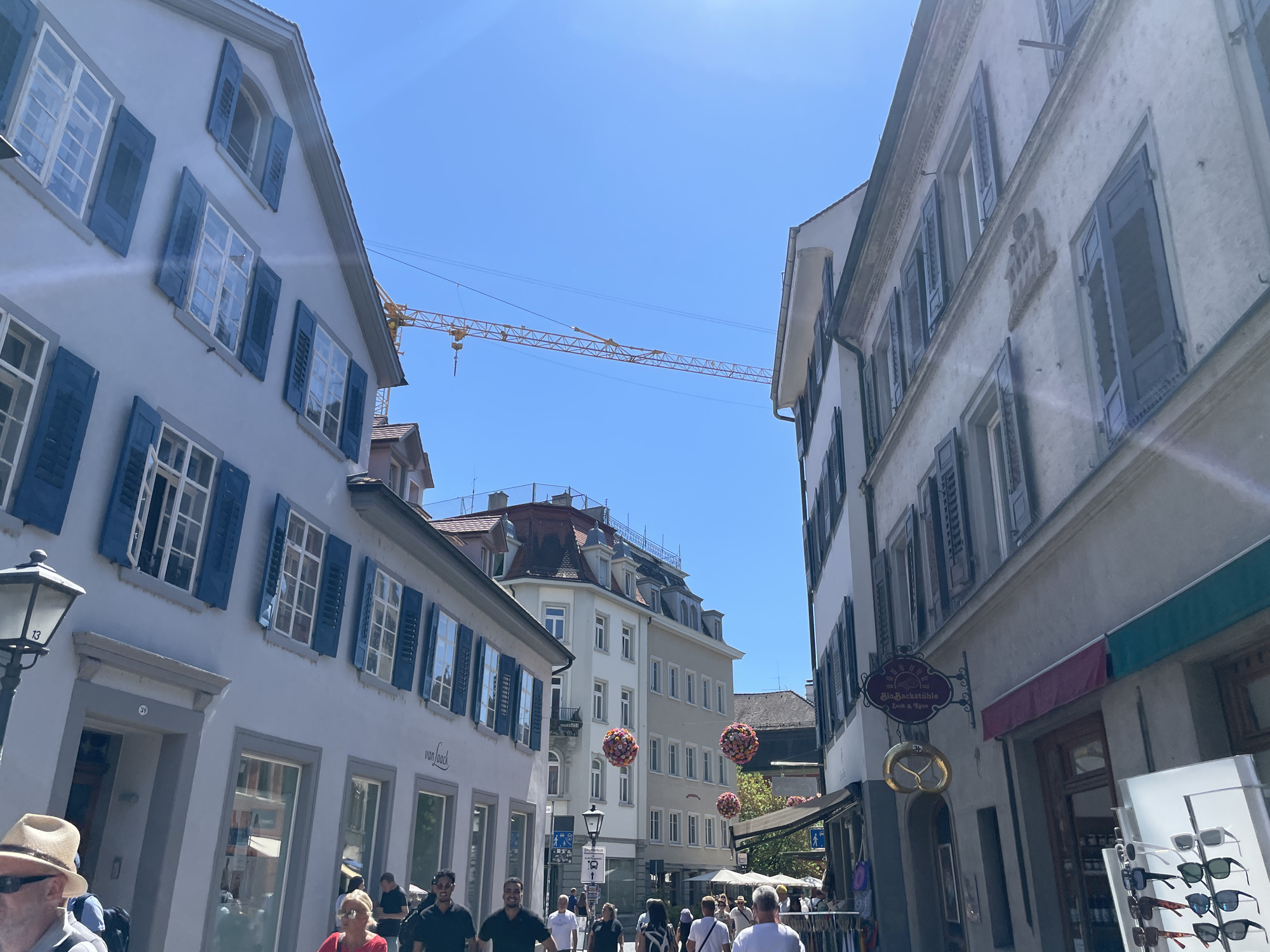 This image captures a street scene in Constance, Germany, characterized by traditional European architecture under a clear blue sky. On the left, a light-colored building features multiple stories with evenly spaced windows adorned with blue shutters, some of which are open. A lamppost with the number "13" is visible on the side of this building. The right side of the street is lined with buildings displaying similar architectural elements, including windows with dark shutters and decorative facades. A yellow construction crane is prominently positioned in the sky, extending across the frame, suggesting ongoing development or renovation in the area. Several people are walking along the cobblestone street, some appearing to be tourists due to their attire. Signs with text in German, such as "van Laack" and "Bio-Backstüble," are visible on the storefronts. Decorative round floral arrangements are suspended in the air between buildings, adding a touch of festivity to the urban environment. The overall impression is a lively, sunny day in a historic European city.