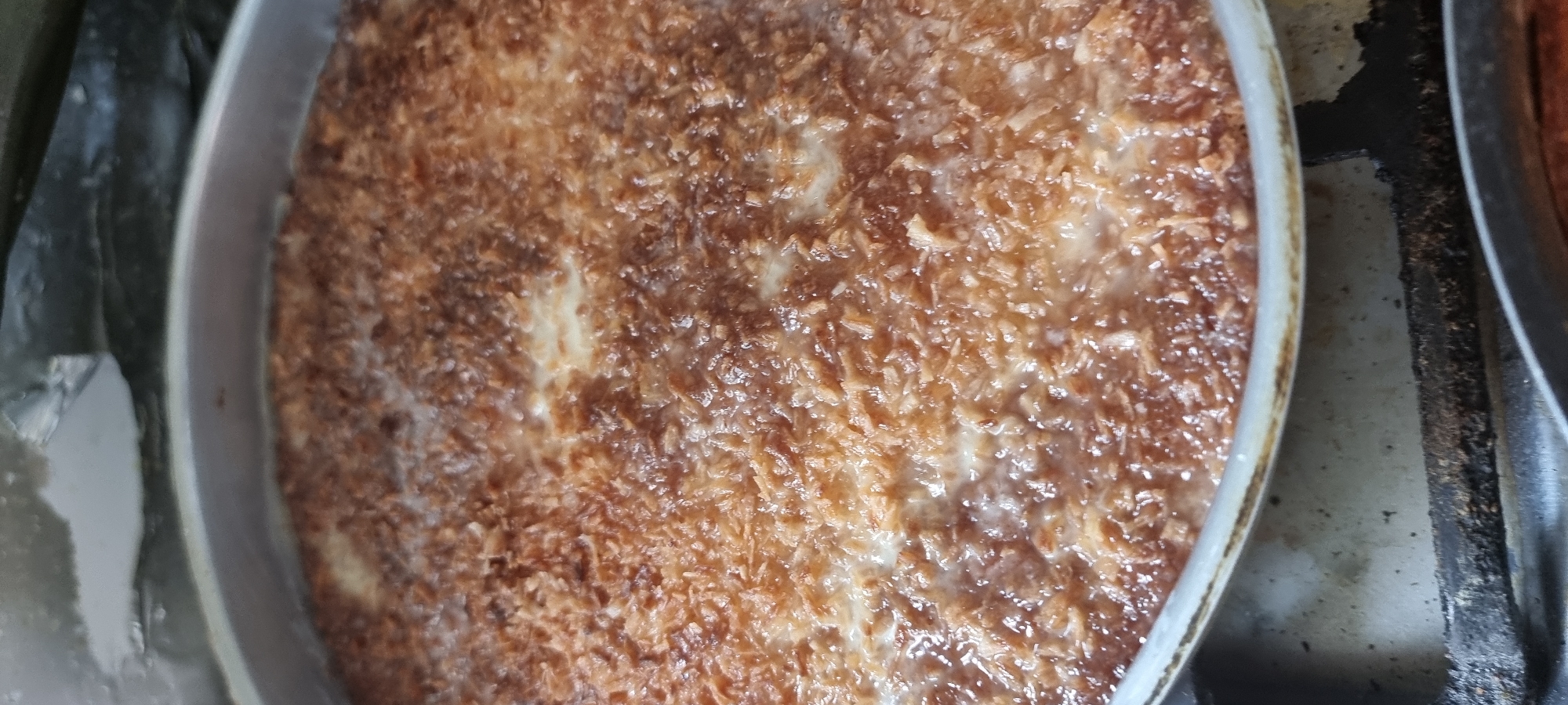 A close-up, overhead shot shows a round, metallic baking pan filled with a dish that appears to be cooked. The surface of the dish is covered in what looks like toasted coconut flakes, giving it a textured, golden-brown appearance. Some areas of the dish appear lighter, suggesting a creamy base underneath the topping. The pan itself is silver-colored with a raised rim. The background is out of focus, but hints at a kitchen environment with dark, possibly metallic surfaces. No people or specific location details are visible. The image was taken in Aktobe, Kazakhstan.