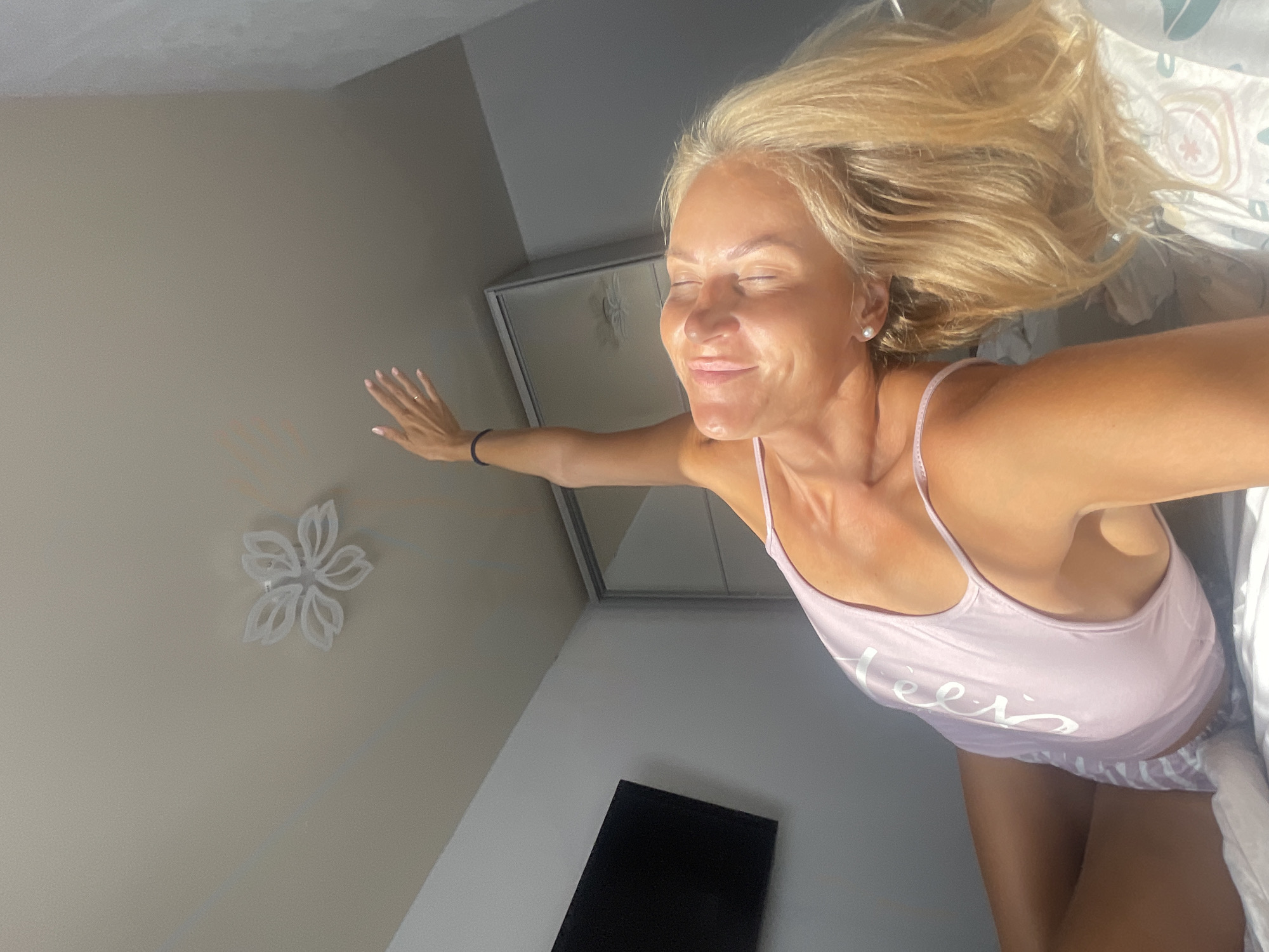 A blonde woman is performing a yoga pose in a bedroom. She is lying on her side with her legs bent and her upper body twisted, with her left arm extended towards the ceiling. She is wearing a light pink tank top with white writing and striped pajama bottoms. The room has white walls and a light-colored ceiling with a flower-shaped light fixture. A flat-screen television is mounted on the wall to the left. To the right, there is a closet with mirrored doors. The woman appears to be bathed in sunlight, suggesting it is daytime. There is no information about the location being in Baranovka, Russia.