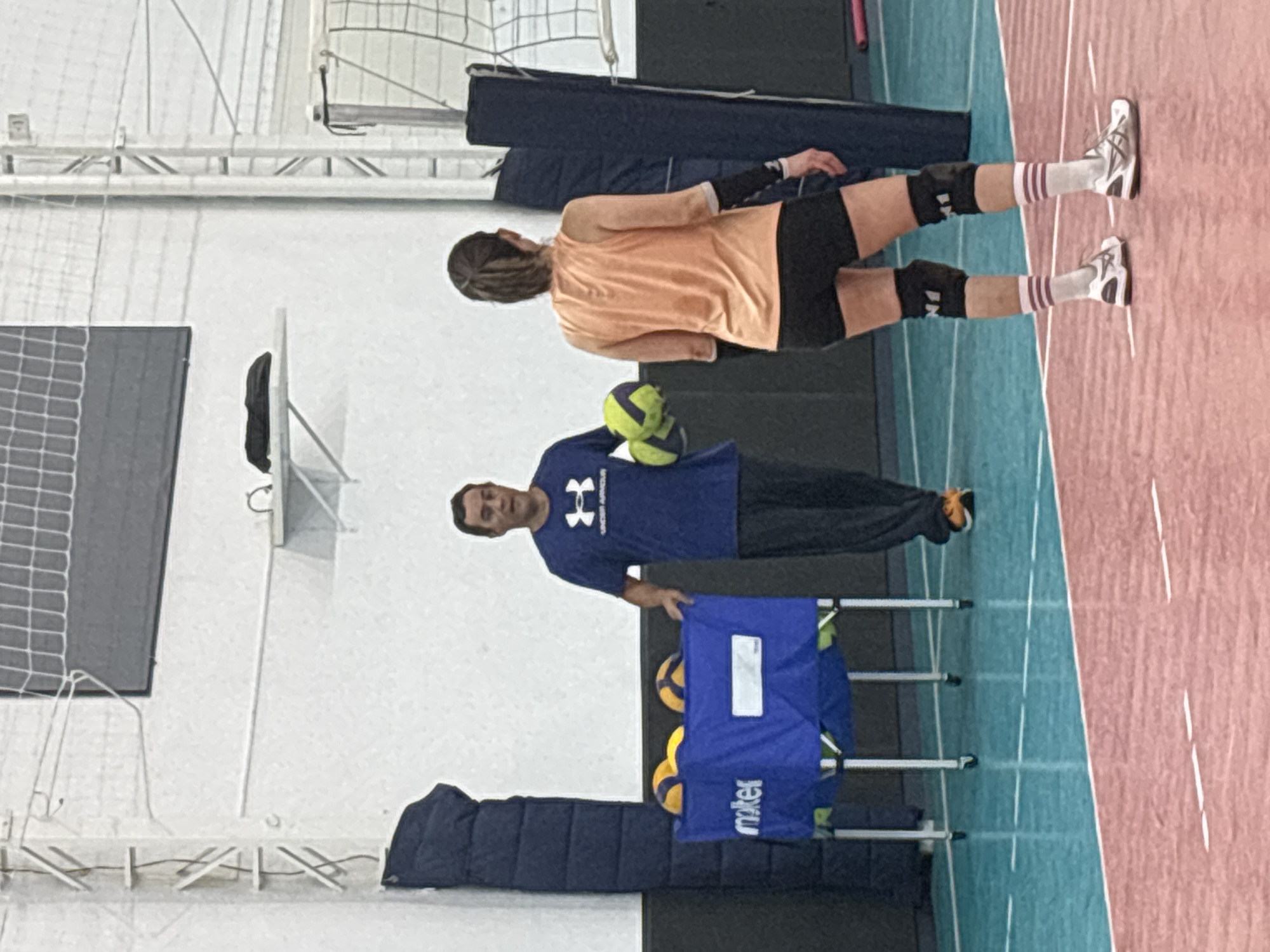 A coach and volleyball player practice indoors, likely in a gymnasium. The coach stands near a ball cart with several volleyballs. He wears a blue Under Armour shirt and black pants. The player is on the other side of the court, wearing an orange shirt, black shorts, knee pads, and striped socks. She appears ready to practice or train. The court is marked with red and blue sections. The location is unknown and cannot be determined as São Paulo, Brazil.