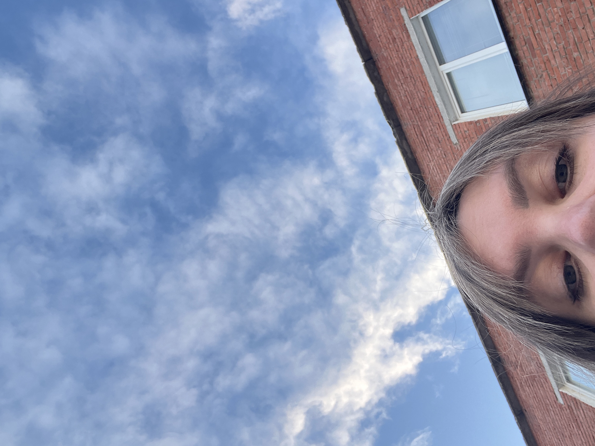 The image shows a low-angle shot looking up at a brick building and a blue sky with scattered clouds. A person's face, specifically their eyes and forehead, is visible in the foreground, cropped from the top. The building appears to be made of red bricks and has white-framed windows. The sky is a bright blue with wispy white clouds. The overall impression is that of a candid outdoor shot, possibly taken while the subject is lying down or looking upwards. The location is identified as Noviy, Russia.