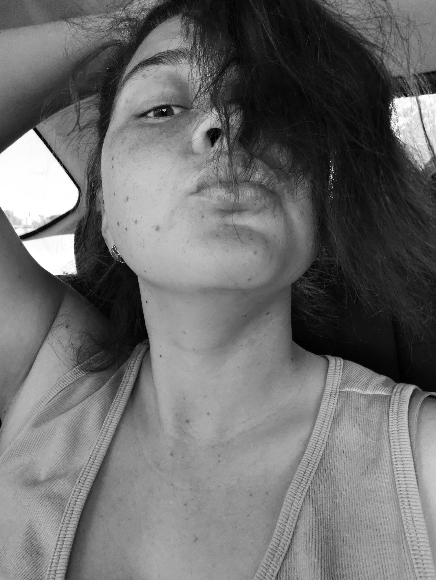 The black and white image depicts a woman with freckles and dark hair partially obscuring her face. She is wearing a tank top and appears to be sitting in a car, with a window visible in the background. Her hair is messy and she has a slight pout on her lips. Her arm is raised, possibly resting on the headrest of the car seat. The woman's location in Chaikovsky, Russia, is not discernable from the image.