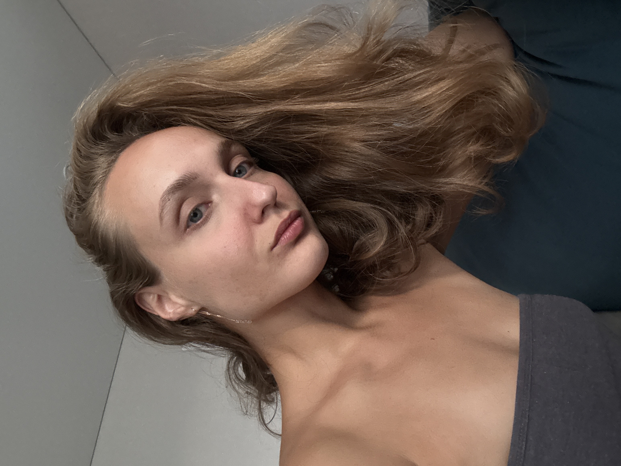 The image shows a close-up, head-and-shoulders shot of a young Caucasian woman with fair skin and long, wavy blonde hair. She is looking downwards and slightly to the left, with her head tilted. Her eyes are blue, and she has natural-looking makeup. She is wearing a grey strapless top. A delicate chain earring with a small pendant is visible in her left ear. The background consists of a plain, light-colored wall and a dark teal surface, possibly a sofa or chair. The lighting is soft and natural, highlighting the texture of her hair and skin. The location is not discernible from the image itself, and there is no information about the city of Подшивалово, Russia.