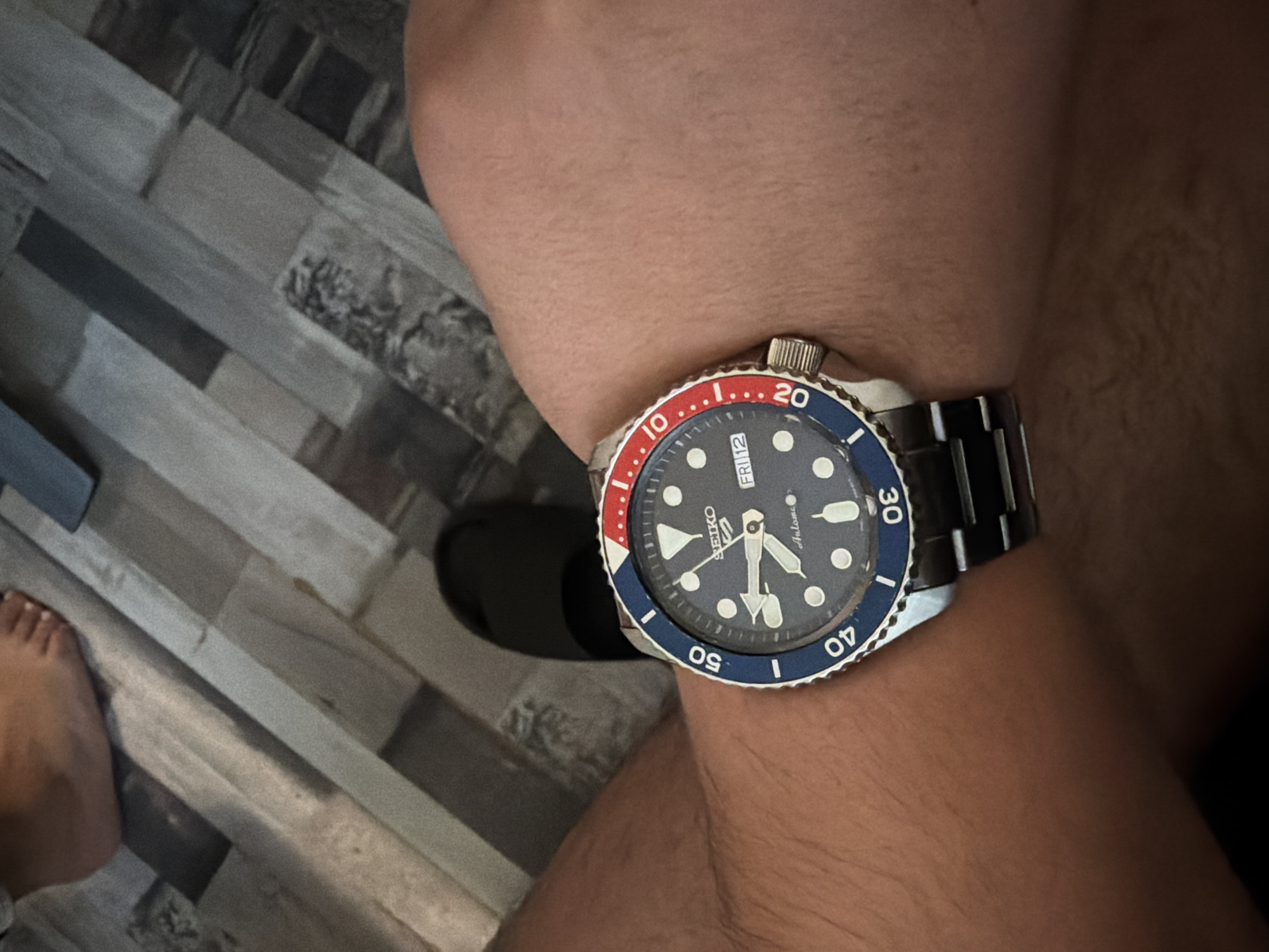 A close-up, high-angle shot shows a person's wrist wearing a Seiko diver's watch. The watch has a dark blue face with white hour markers and hands, and a red and blue bezel. The date and day are visible as "FRI 12". The watch band is made of metal. The person's skin appears to be light-skinned. In the background, a floor with a wood-grain pattern is visible, with some light and dark planks. A sliver of a bare foot is visible at the top left of the frame. The scene is indoors and appears to be a casual setting. There is no information in the image to determine the location within Sofia, Bulgaria.