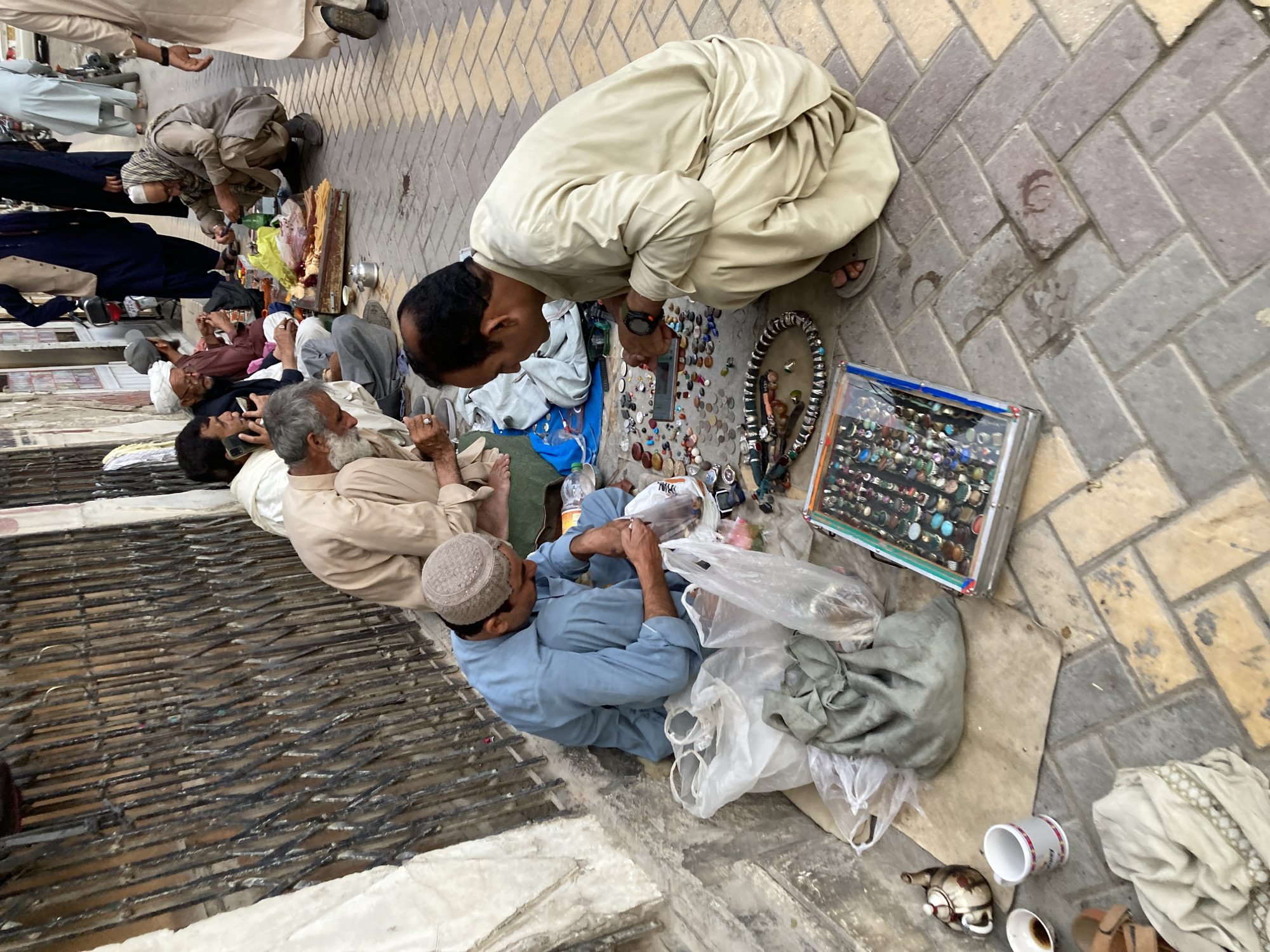 This image captures a bustling street scene in Quetta City Tehsil, Pakistan, featuring several men engaged in commerce and social interaction. In the foreground, a man in a light beige shalwar kameez squats, attentively examining a street vendor's display of small items spread on a cloth. The merchandise includes numerous colorful gemstones, metal coins, and rings housed in a clear plastic case. A large beaded necklace is also part of the display.
To his left, another man, wearing a light blue shalwar kameez and a prayer cap, sits on the ground, appearing to adjust items in a plastic bag. Nearby, an older man with a white beard is conversing on a mobile phone, while further back, another individual looks at his device. The ground consists of herringbone-patterned paving stones, interspersed with dirt patches and a metal grate. Personal effects such as plastic bags, a teapot, and white cups (one containing a dark liquid) are placed on the ground.
The informal street market setting suggests daytime activity under clear, dry weather conditions. The traditional attire of the men and the nature of the vending activity are characteristic of the region. No legible text is visible within the frame. The overall atmosphere conveys a sense of casual engagement and daily life on a busy street.
