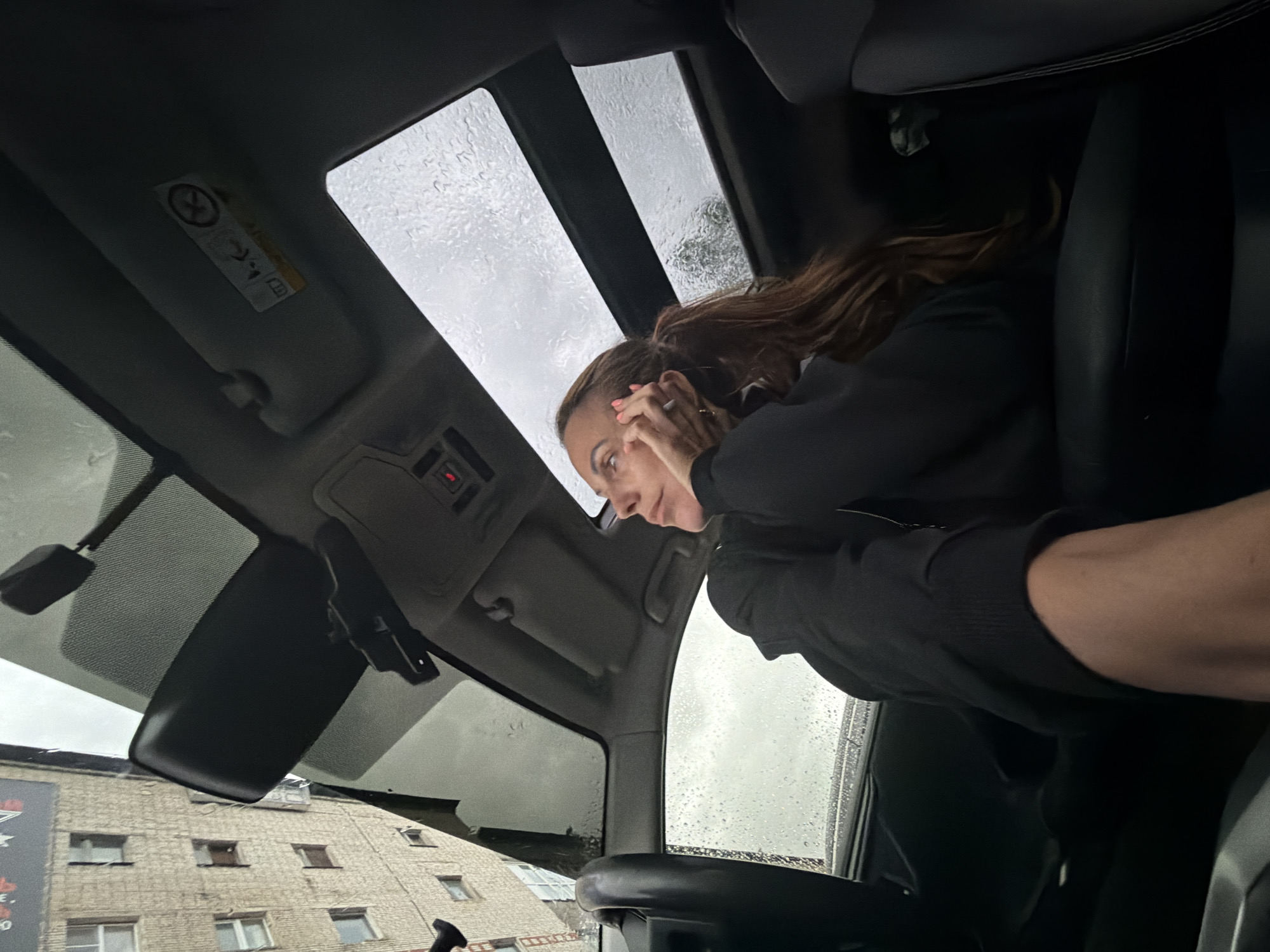 A woman with long, brown hair is looking downwards inside a car. She is wearing a black jacket and has her hands near her face. The car's interior is visible, including the sunroof which shows a rainy sky with water droplets on the glass. The view from the car's windows reveals a brick building with multiple windows, suggesting an urban or suburban setting. The time of day appears to be overcast, likely daytime given the light. The overall mood seems neutral or perhaps slightly contemplative, as indicated by the woman's posture. The image is captured from a low angle, looking upwards towards the car's roof and the woman. There is a visible warning label with symbols on the car's sun visor.