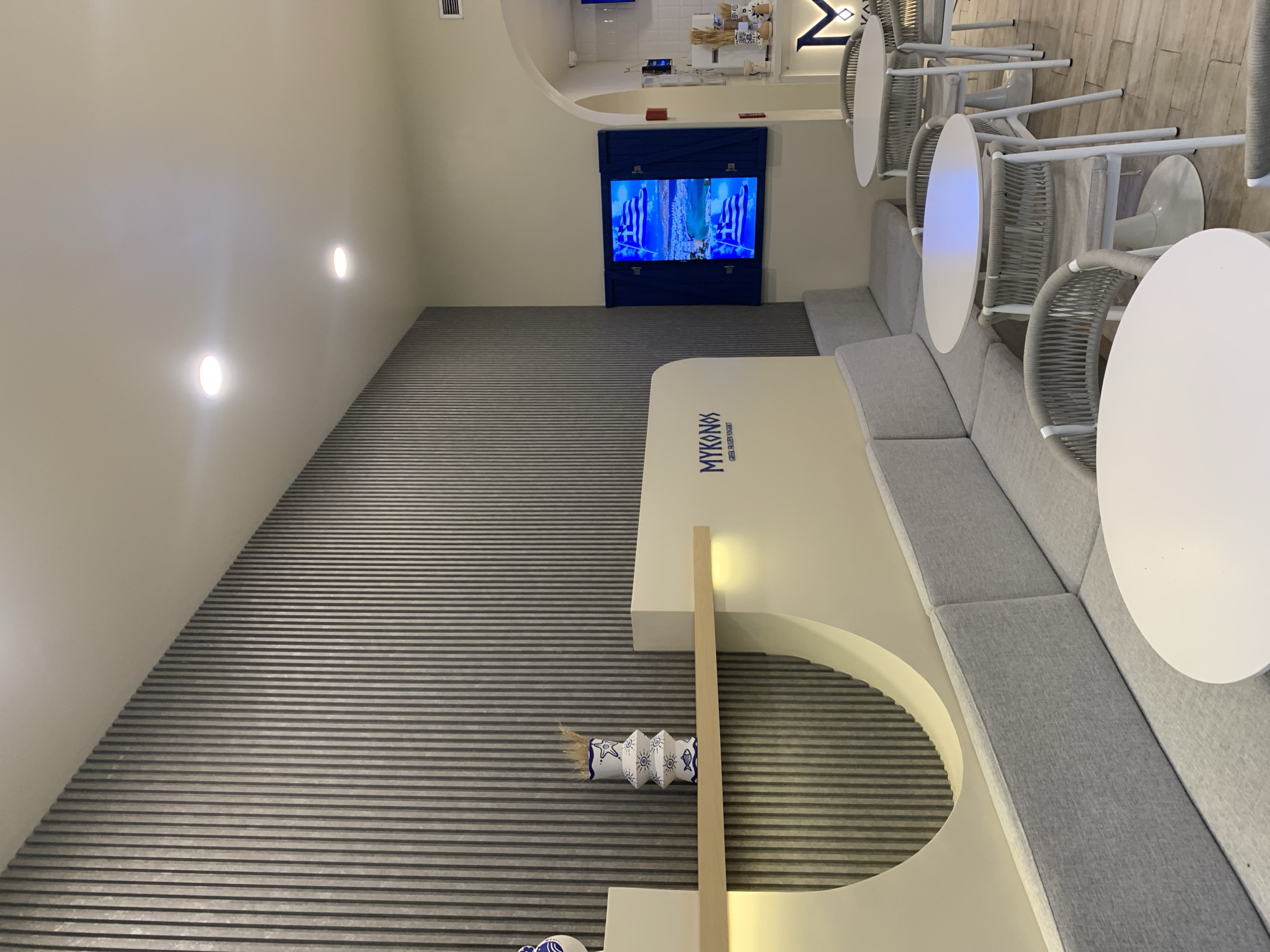 This media file captures the interior of a modern, Greek-themed cafe or restaurant named "Mykonos Greek Street Food" in Pampatar, Venezuela. The space features a clean, minimalist design dominated by white, light grey, and blue tones.
On the right side of the image, there is a seating area with a long, built-in sofa upholstered in light grey fabric, curving along a white partition. In front of the sofa are several round, white tables accompanied by chairs with light grey woven seats and white metal frames. The floor beneath the tables is light-colored, possibly tiled or wood-patterned.
The left side of the room features a textured wall with horizontal grey slats or ribs, possibly for acoustics. Embedded in this wall is a decorative niche or cutout. A television screen, encased in a blue framed structure, is mounted on a wall towards the back, displaying an image with prominent blue and white patterns, resembling the Greek flag. Above the TV, and further into the background, a large blue "M" logo is visible.
Recessed circular lights illuminate the white ceiling and upper walls. On the textured wall, near the seating area, there's a decorative rolled paper object with blue patterns (stars, fish, suns) and a natural fiber tassel, likely contributing to the Mediterranean ambiance. The main partition features the "MYKONOS GREEK STREET FOOD" logo in blue lettering. The entire setting appears bright and inviting, suggesting a contemporary eatery. No people are visible in the scene.