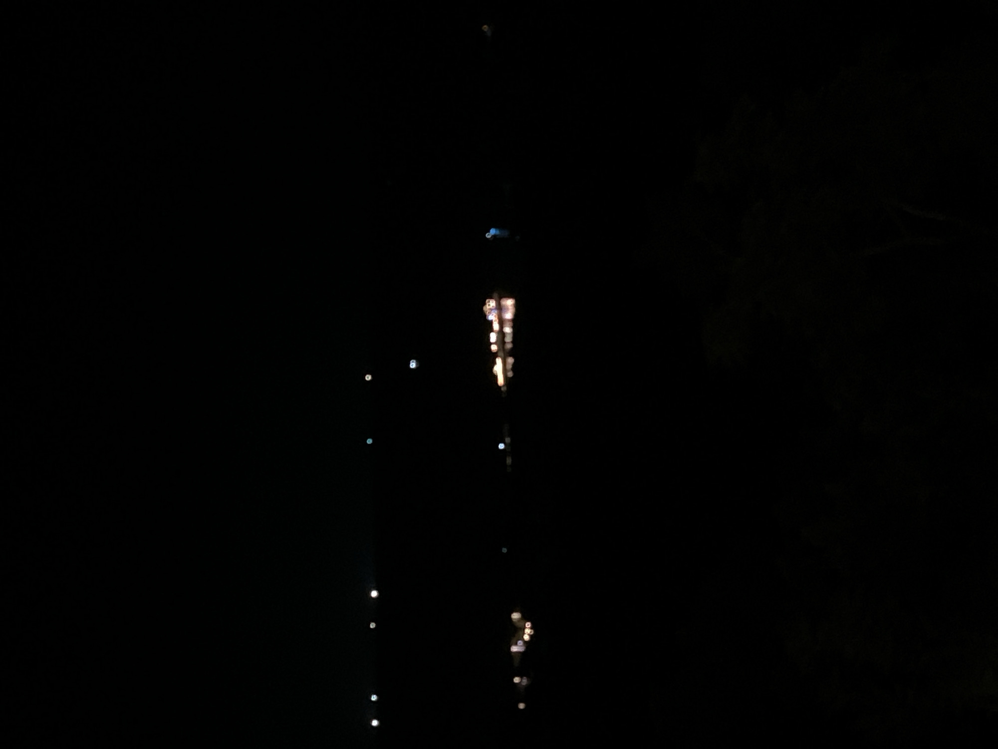 The image shows a dark night scene with several blurred lights reflecting on what appears to be water. There are some scattered lights in the background that suggest distant structures or boats. One cluster of lights in the mid-ground appears to be a boat or a series of boats on the water, with reflections shimmering below. Another blurred light, possibly from a buoy or small vessel, is visible further to the right. The overall impression is one of a quiet night by the water, likely in a coastal area. Given the prompt's specificity, this scene could be located along the coastline of Karimunjawa, Indonesia, known for its numerous islands and coastal activities.