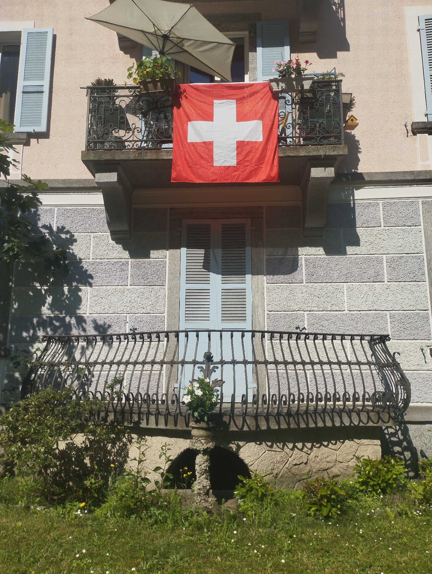 A Swiss flag hangs from a balcony of a light pink building. The flag is red with a white cross. The balcony is ornate and made of black wrought iron. There are potted plants and an umbrella on the balcony. Below the first balcony, there is a second, larger balcony, also with a black wrought iron railing. A door with blue shutters is visible behind the second balcony. The building's facade is textured with light-colored stone blocks. In front of the building, there is a grassy area with several small white flowers and green bushes. The scene is illuminated by sunlight, casting shadows on the building and the ground.