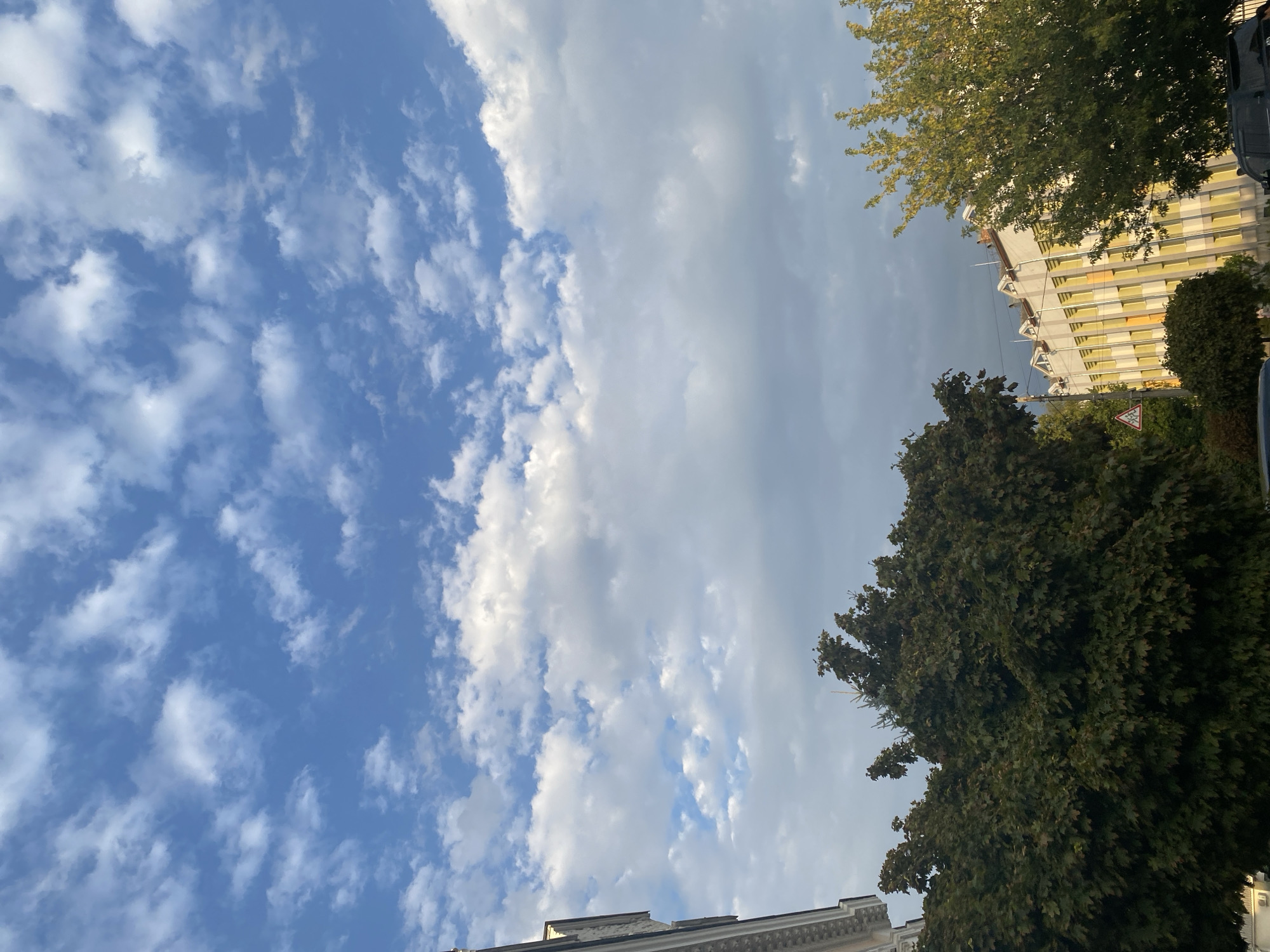 A low-angle, vertical shot captures a cloudy sky over buildings and trees in an urban setting. The sky is a bright blue with scattered white, puffy clouds, some of which appear thin and wispy. On the left side of the frame, a portion of a building's facade is visible, featuring white decorative molding along its edge. In the foreground, a large, dense tree with dark green leaves dominates the left side. To the right, another tree with lighter green foliage stands, partially obscuring a building with a light-colored facade and yellow window frames. The building has a distinctive triangular architectural detail along its roofline. A traffic sign is visible in the middle distance. The overall impression is of a partially overcast day in a city.