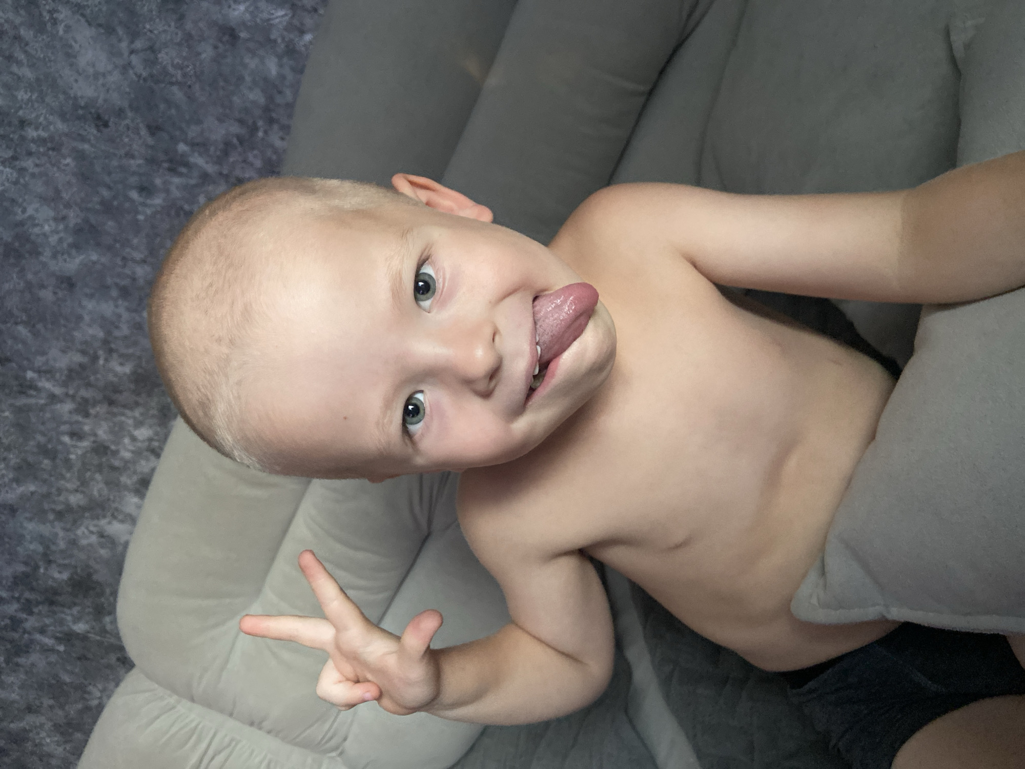 A young, bare-chested boy with a shaved head and blue eyes is shown in a close-up shot, sticking his tongue out and holding up two fingers in a peace sign. He is lying on a light-colored couch with a textured gray background. The boy's expression is playful. The image provides no information about the location in Prokopyevsk, Russia, or any other specific geographical details.