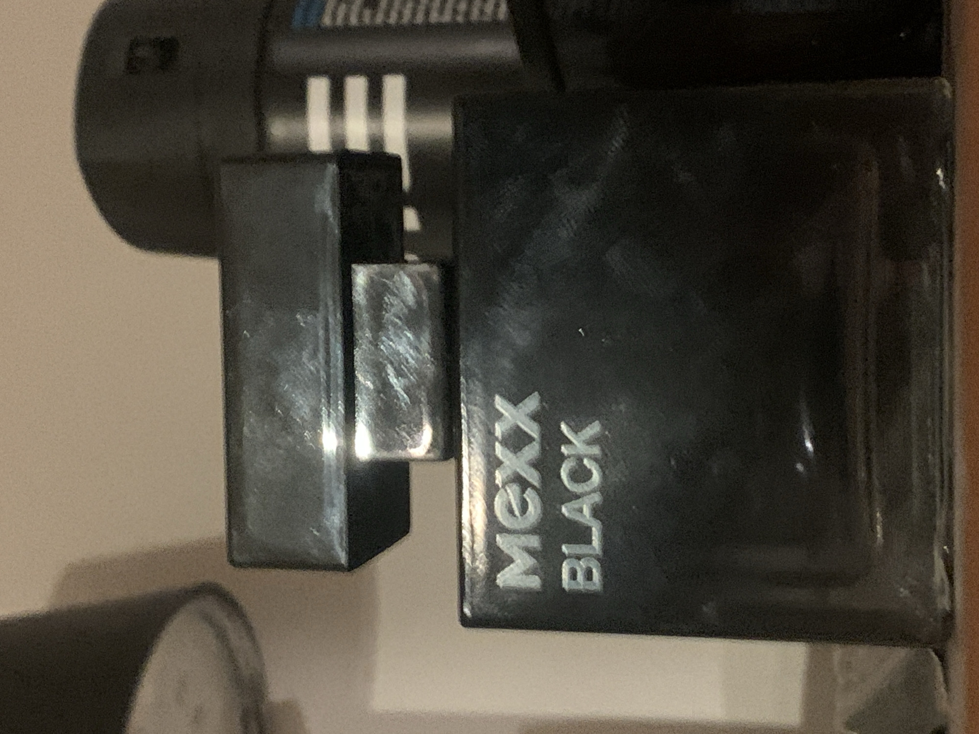 The image displays an arrangement of dark-colored personal care products, possibly fragrances, set against a light background. The primary subject is a rectangular black bottle with a dark, metallic-looking cap. The cap is detached and positioned to the left of the bottle, partially obscuring the top left corner of the main bottle.
The most prominent text, "MEXX BLACK," is visible vertically in silver or white lettering on the front of the black bottle. Behind these items, a larger, dark cylindrical object is partially visible. This background object features several distinct white horizontal stripes and some faint, dark text at its top, where "F P" can be vaguely discerned.
The items are situated on a flat, light-colored surface, which creates visible shadows, suggesting an indoor setting with directed lighting. The background behind the objects is also light-colored. The image has a vertical orientation. There are no people or specific activities depicted; it is a still-life arrangement of products. The surfaces of the bottles show some reflections and minor smudges or dust particles.