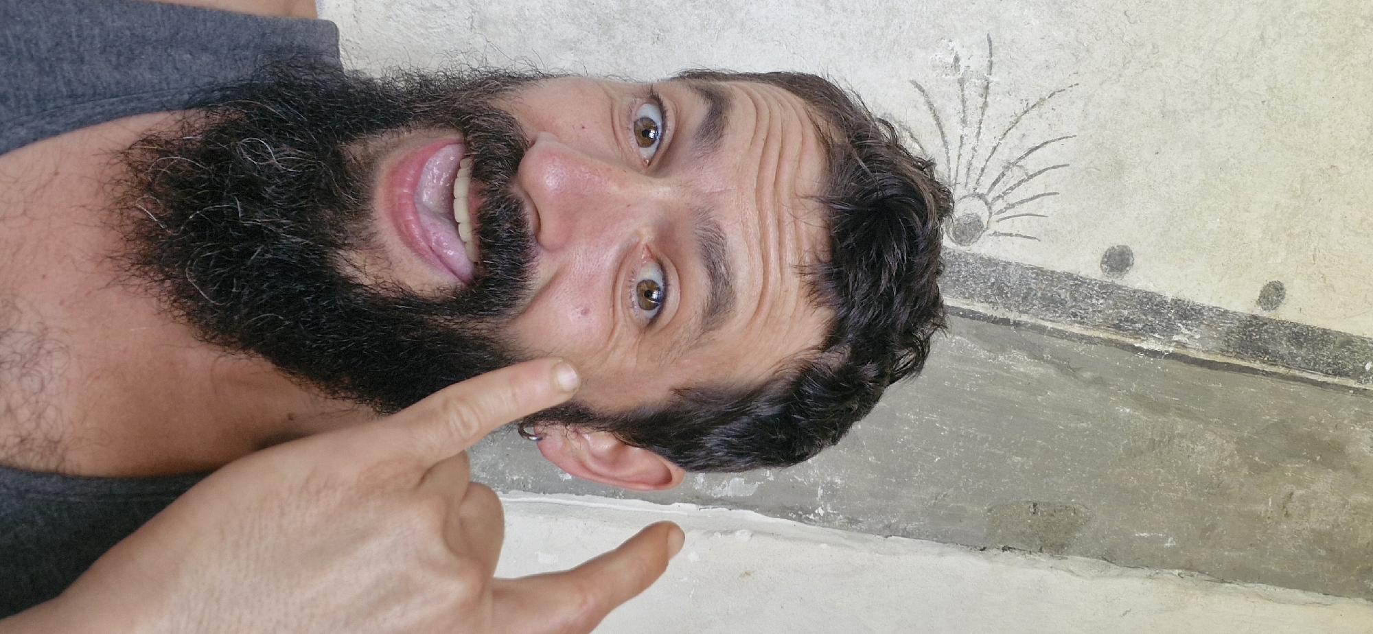 In a close-up, waist-up shot, a Caucasian male with brown hair, a full beard, and light brown eyes is pictured against a textured, off-white wall with faint gray markings. He is wearing a gray tank top. The man is looking directly at the camera with his mouth open, tongue sticking out, and a playful expression. His right index finger is extended, pointing towards his cheek, and his left hand is out of frame. The lighting is natural, casting soft shadows. The background wall has a distinct vertical, textured strip of concrete or plaster. The image is taken outdoors, possibly on a wall of a building.