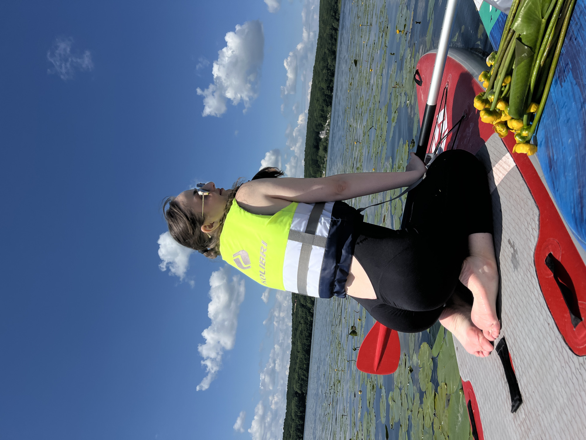 A woman kneels on a paddleboard in a lake, facing away from the camera and looking to her right. She wears a bright yellow life vest with reflective stripes and black leggings. Her feet are bare and visible on the paddleboard. A red paddle rests on the left side of the board, and a bunch of yellow water lily flowers and leaves are on the right. The lake is covered with lily pads, and the background shows a dense, green forest line under a bright blue sky with scattered white clouds. The location is identified as the city of Bila, Ukraine.