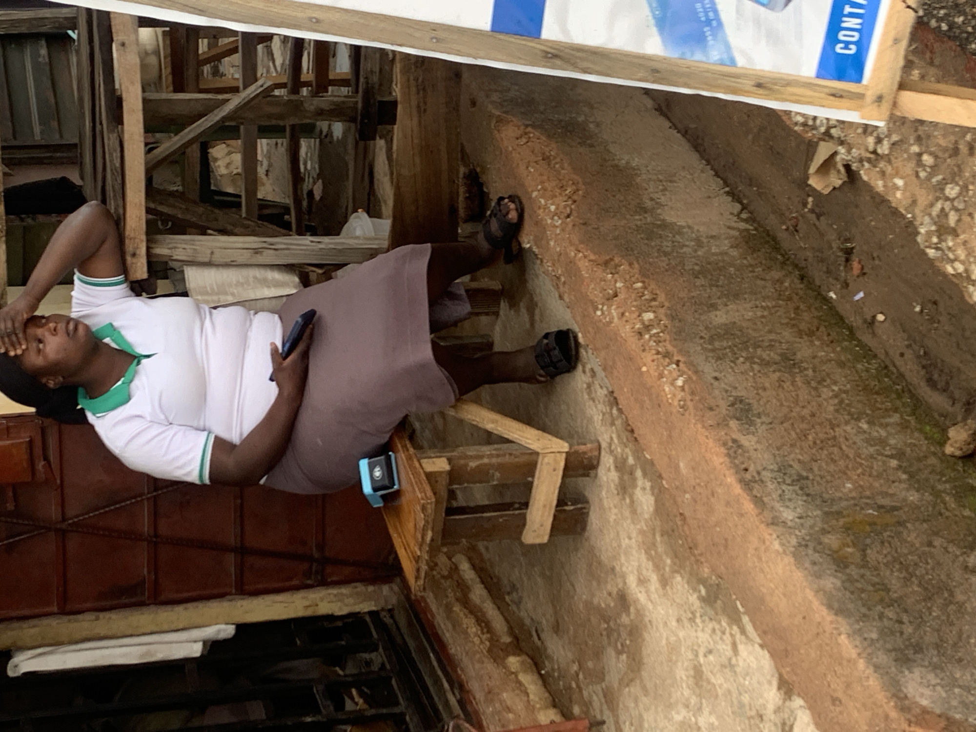 A woman in a white t-shirt with green trim and a brown skirt is sitting on a weathered wooden chair in an outdoor area. She is holding a phone in her left hand and touching her forehead with her right hand, appearing stressed or thoughtful. Her footwear is dark-colored sandals. The location appears to be a commercial or semi-industrial outdoor space, potentially in Jos, Nigeria, based on likely context.
The background includes a jumble of wooden structures, metal bars, and weathered concrete, suggesting a bustling, somewhat disorganized environment. A sign with the word "CONTA" is partially visible above her. The area looks aged and worn with visible signs of weather exposure. A drain is visible on the right side.