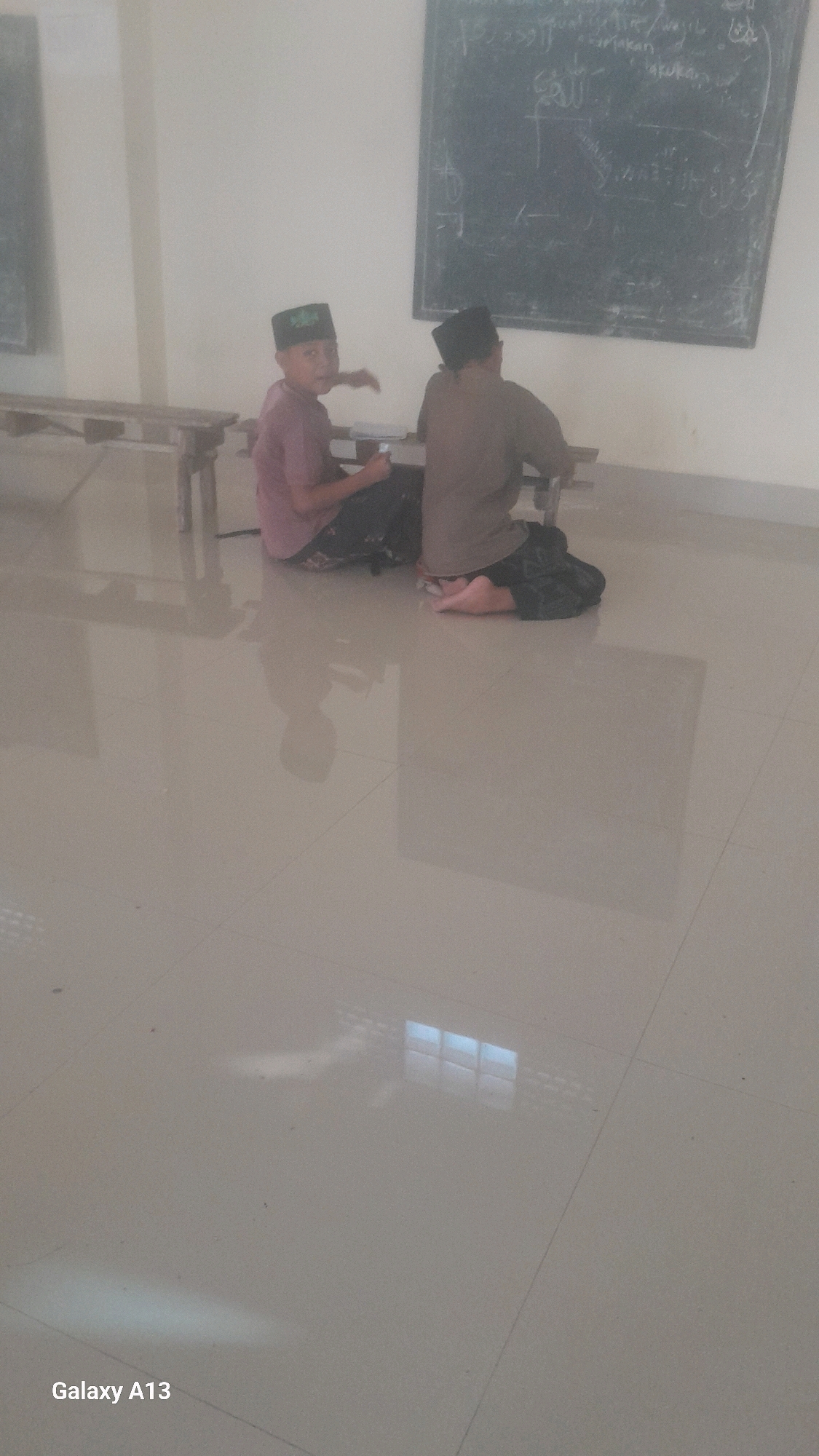 Two young boys are sitting on the floor in front of a chalkboard. The boy on the left is wearing a pink shirt and a green kufi, and he is looking at the camera. The boy on the right is wearing a brown shirt and a black kufi, and he is looking at the chalkboard. The chalkboard has Arabic writing on it. The floor is tiled and reflective. The image was taken in a classroom in Ngelo, Indonesia.