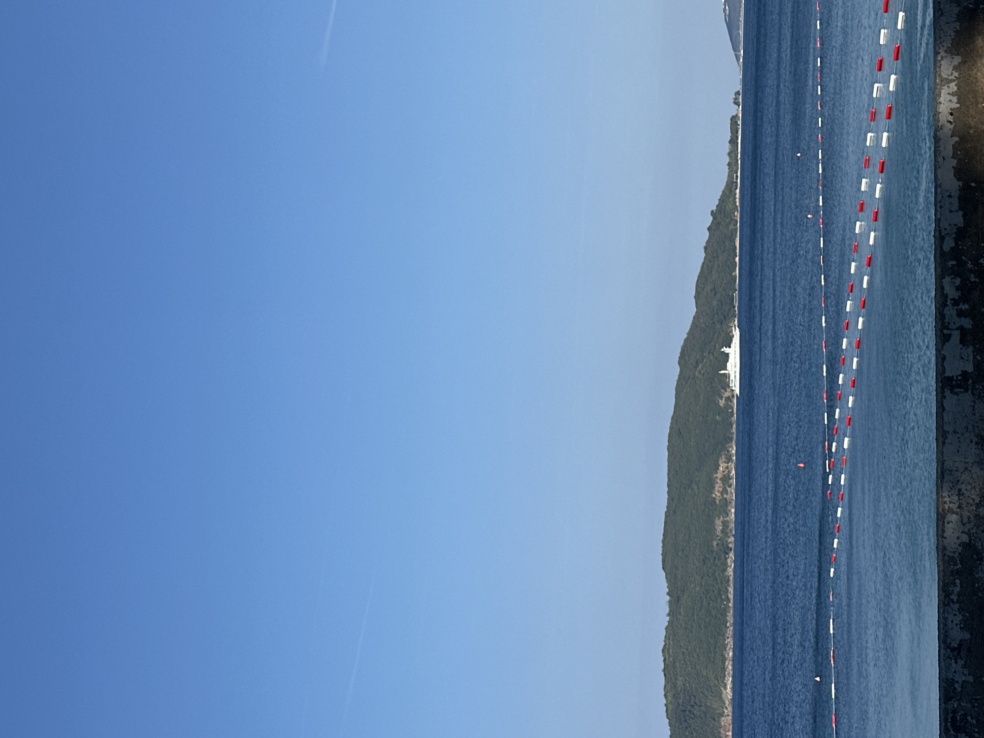 A wide, clear blue sky dominates the upper portion of the image. Below, a calm, dark blue sea stretches across the middle ground. In the distance, a lush green, treed island or peninsula forms a hilly coastline. A white cruise ship is visible at anchor off the coast of the island. In the foreground, a rope barrier with alternating red and white buoys divides the water, creating a designated swimming area. A stone wall is present at the very bottom of the image, bordering the water. The scene appears to be a coastal area, likely a beach or harbor, in Boreti, Montenegro, given the typical landscape and the presence of buoys marking a swimming zone. There are no people visible in the image.