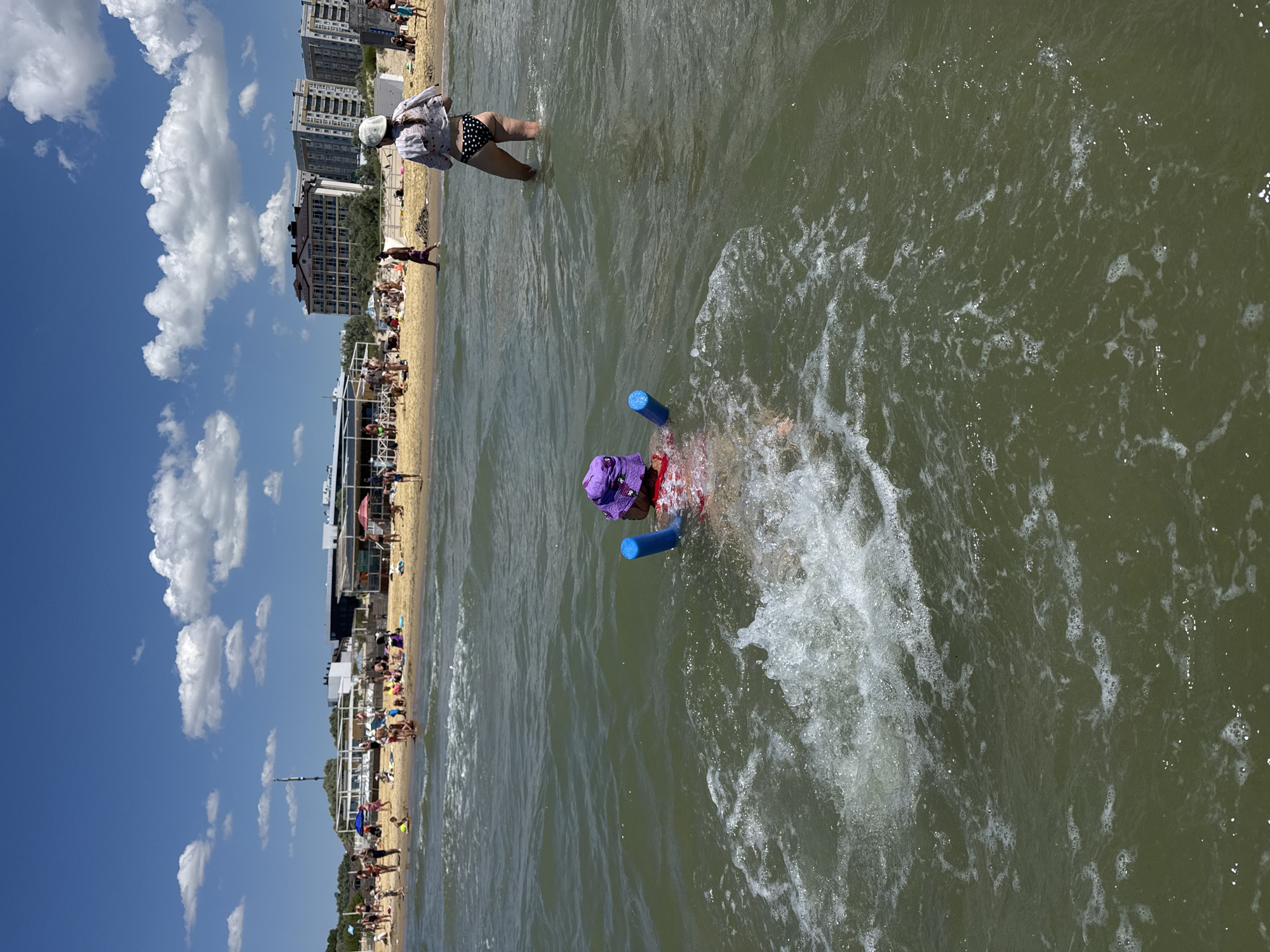 The image depicts a sunny day at a crowded beach in Prostorny, Russia. In the foreground, a child wearing a purple hat and a red swimsuit is playing in the water, holding onto two blue pool noodles. The child is splashing and creating ripples. Further back, on the sandy beach, numerous people are visible, some are sunbathing, others are walking or sitting. A few beach umbrellas and structures are present along the coastline. In the background, apartment buildings and trees line the horizon under a blue sky with scattered white clouds. The sea is a murky green with small waves.