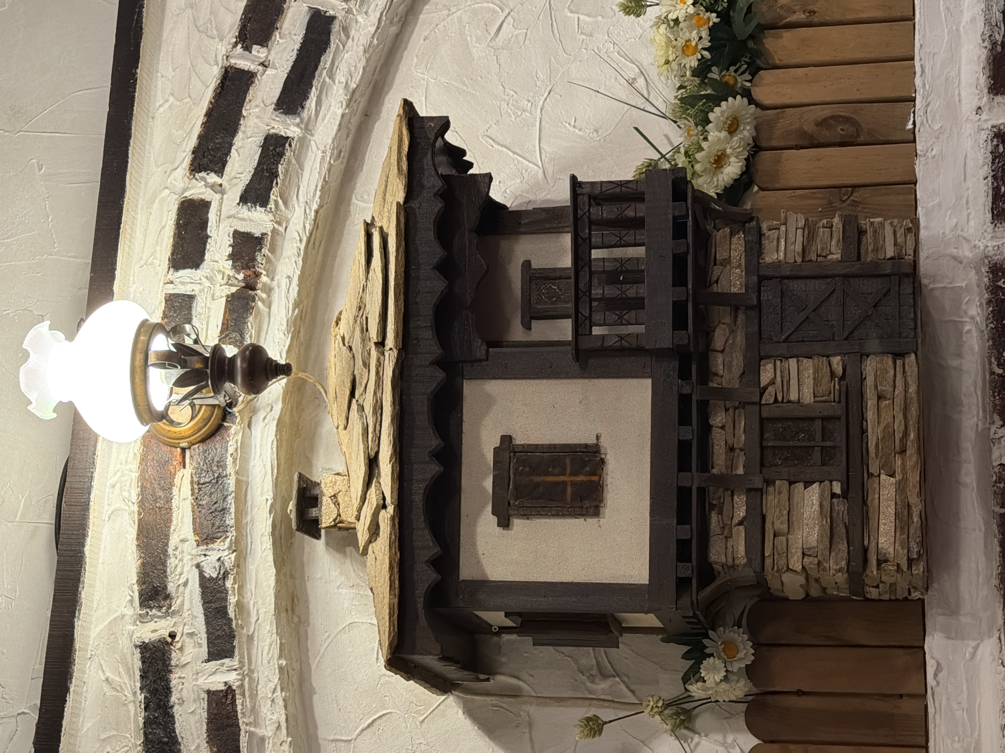 A wall-mounted decoration resembling a miniature, multi-tiered house is affixed to a textured white wall. The house incorporates various materials including wood, stone, and possibly plaster. Decorative flowers adorn the wall around the house, both above and below. To the left, a light fixture with a frosted glass shade is also mounted on the wall. The wall has a faux brick pattern extending from the light fixture upward in an arch. The location is likely a restaurant or bar in Varna, Bulgaria, called "Pivovarna Varna."