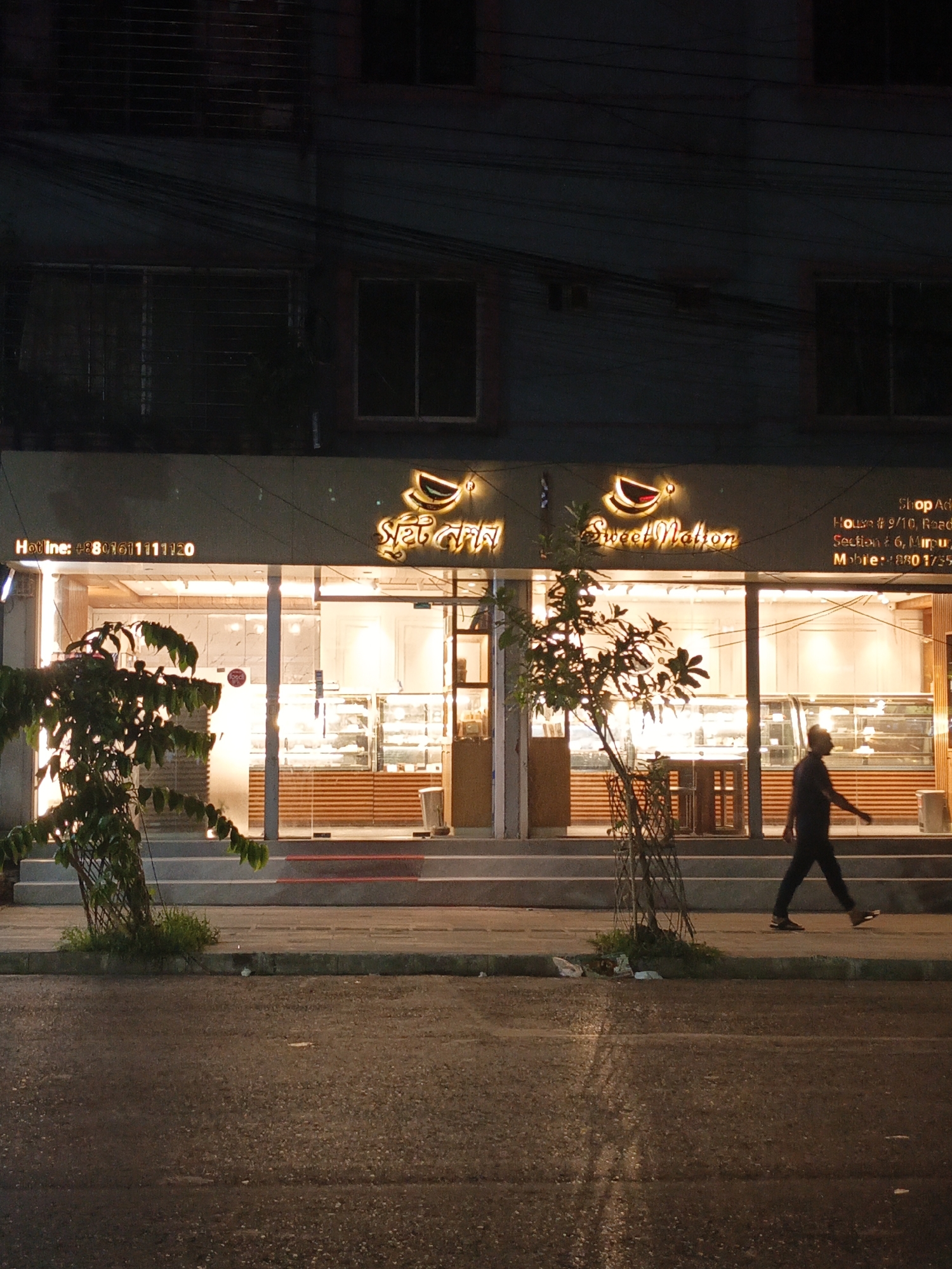 The image shows the storefront of a shop called "Sweet Nation" at night. A man is walking on the sidewalk in front of the shop. The shop address is House # 9/10, Road # 6, Mirpur. The shop is well-lit and displays various baked goods in display cases. There are young trees planted on the sidewalk in front of the store. A wet asphalt road is visible in the foreground. Electrical wires are strung overhead.
