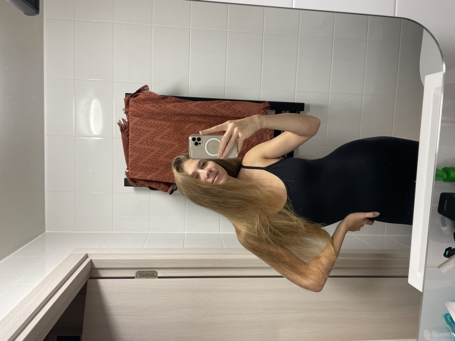 A woman with long blonde hair wears a black dress, posing for a mirror selfie in a bathroom. She holds her phone up with her right hand, and the phone's reflection is visible in the mirror. The bathroom features white tile walls, a towel rack with a brown towel, a vanity, and a large mirror. The woman is smiling at the camera.