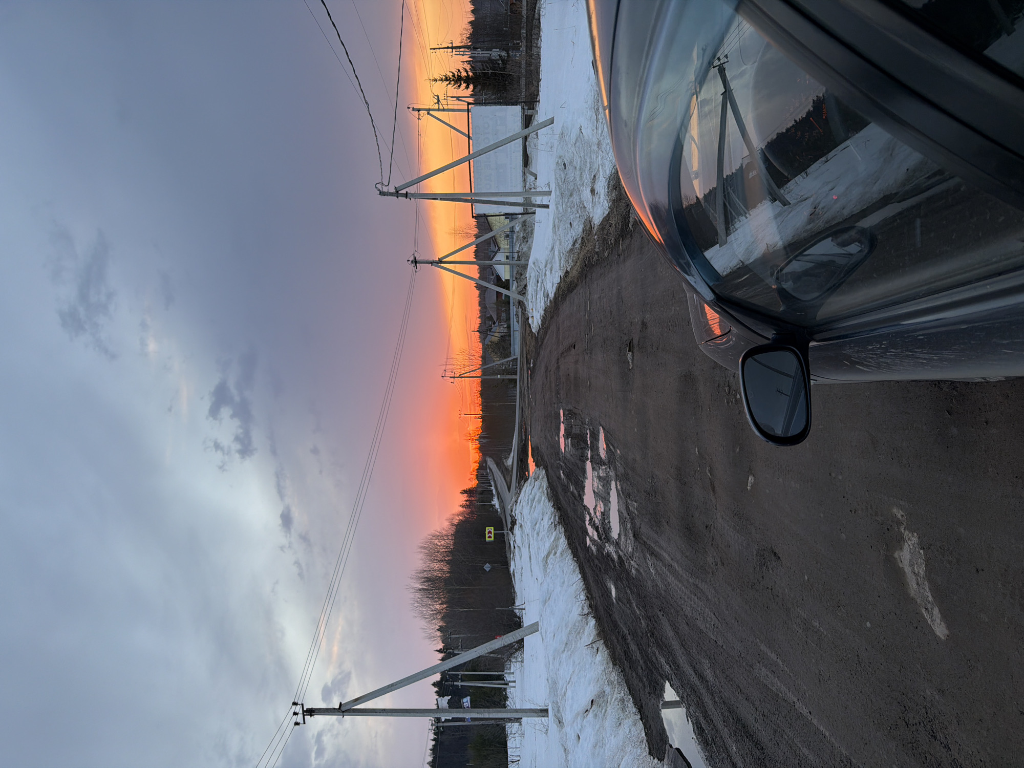 The image shows a dirt road at sunset in a rural area near Izhevsk, Russia. Patches of melting snow are visible along the side of the road. Power lines run along the road supported by wooden poles. A car is partially visible on the right side of the image. The sky is a mix of gray clouds and orange sunset colors. Puddles of water are present on the road surface. A road sign is visible on the left side of the road.