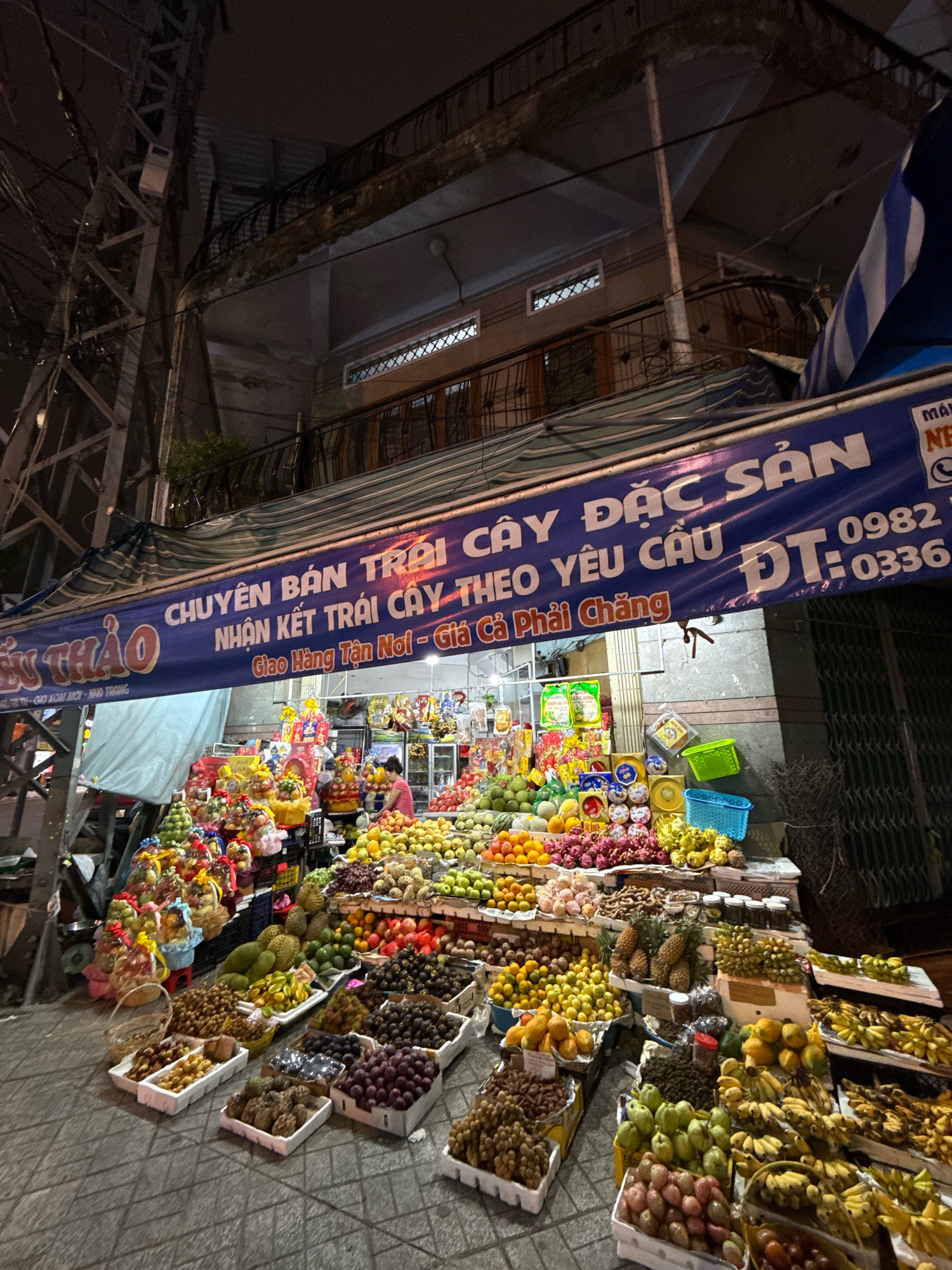 This image captures a bustling night fruit stall in Nha Trang, Vietnam. The stall is brightly lit, with a large blue banner overhead displaying "CHUYÊN BÁN TRÁI CÂY ĐẶC SẢN", translating to "Specialty Fruit Sales". Below this, it reads "NHẬN KẾT TRÁI CÂY THEO YÊU CẦU" and "Giao Hàng Tận Nơi - Giá Phải Chăng", meaning "Accept Fruit Arrangements by Request" and "Home Delivery - Reasonable Prices". The banner also includes a phone number, "0982 0336".
The stall is laden with a wide variety of fresh fruits, arranged in baskets, boxes, and on tiered displays. Visible fruits include durian, bananas, pineapples, longans, grapes, guavas, mangoes, oranges, and dragon fruit, among others. Some fruits are packaged in gift baskets, suggesting they are prepared for sale as gifts. A woman in a pink shirt can be seen working behind the counter, attending to the fruit displays. The stall is situated on a paved street, with a metal structure and power lines visible in the background, indicative of an urban environment. The overall scene depicts a vibrant and active night market scene in the city.