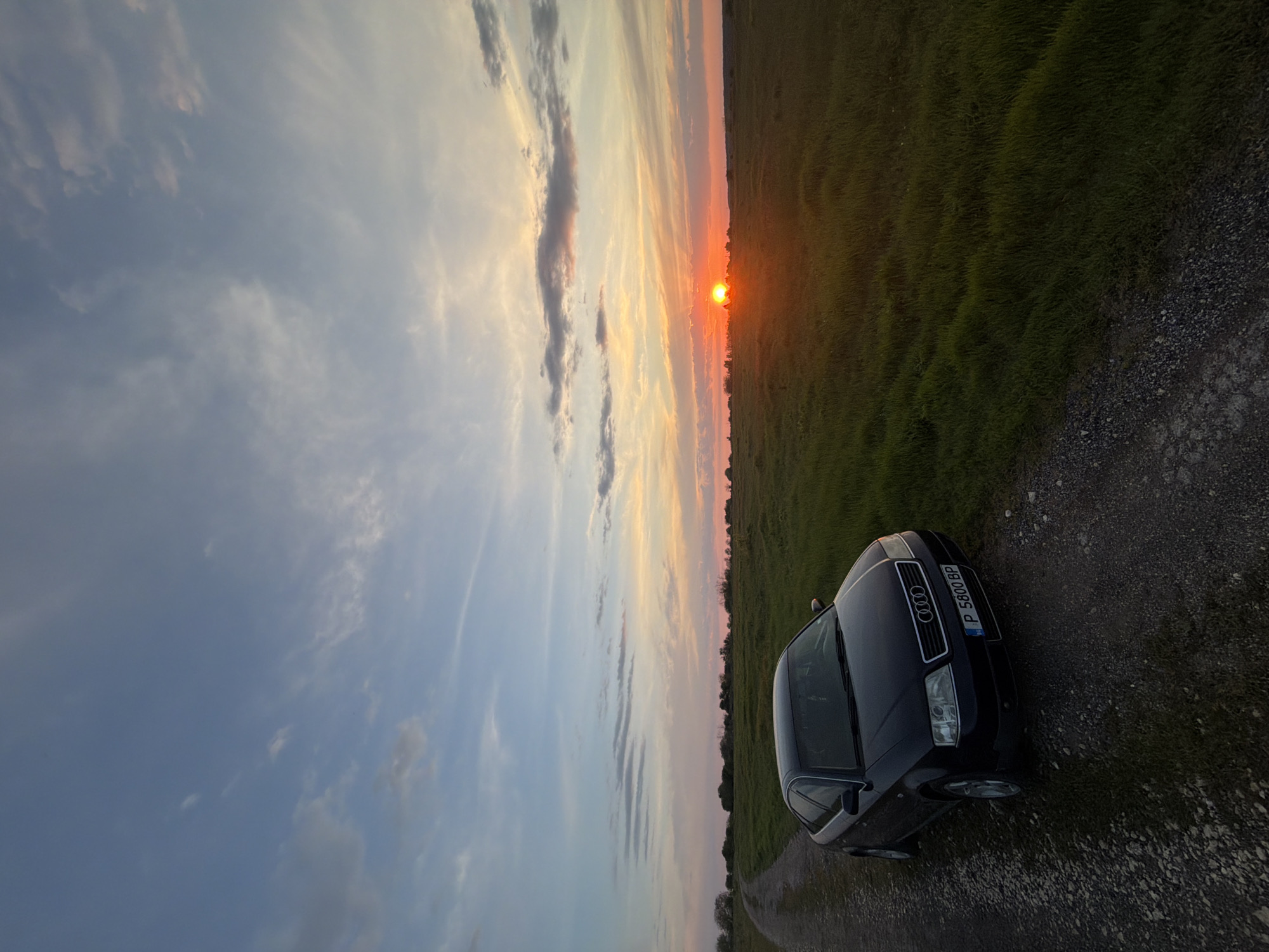 A photograph captures a dark-colored Audi sedan parked on a gravel road beside a field. The sky is visible with a setting sun. The location is Unknown, Unknown.