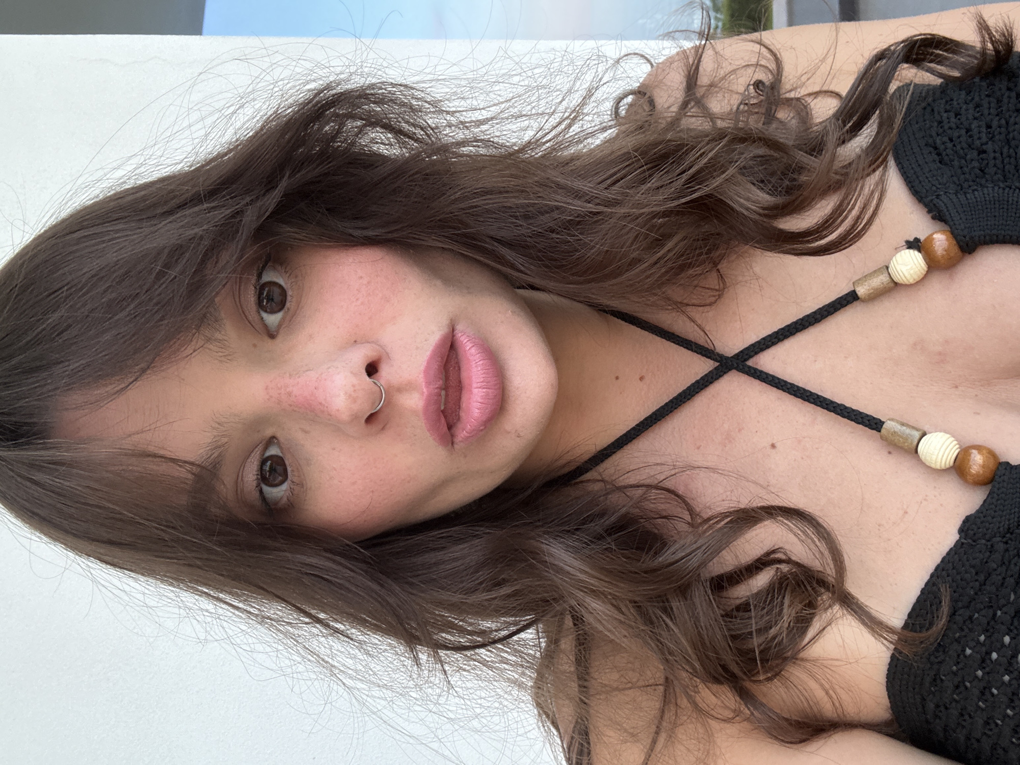 A close-up, slightly angled portrait of a woman with long, wavy brown hair. She has fair skin with visible freckles on her cheeks. A silver nose ring is visible in her left nostril. She is wearing pink lipstick and has brown eyes. She appears to be wearing a black crocheted top with a decorative string with wooden beads across the chest. The background is blurry, showing a white wall and a hint of blue sky, suggesting she may be outside. The location, Siracusa, Italy, cannot be verified solely from the visual content of the image.