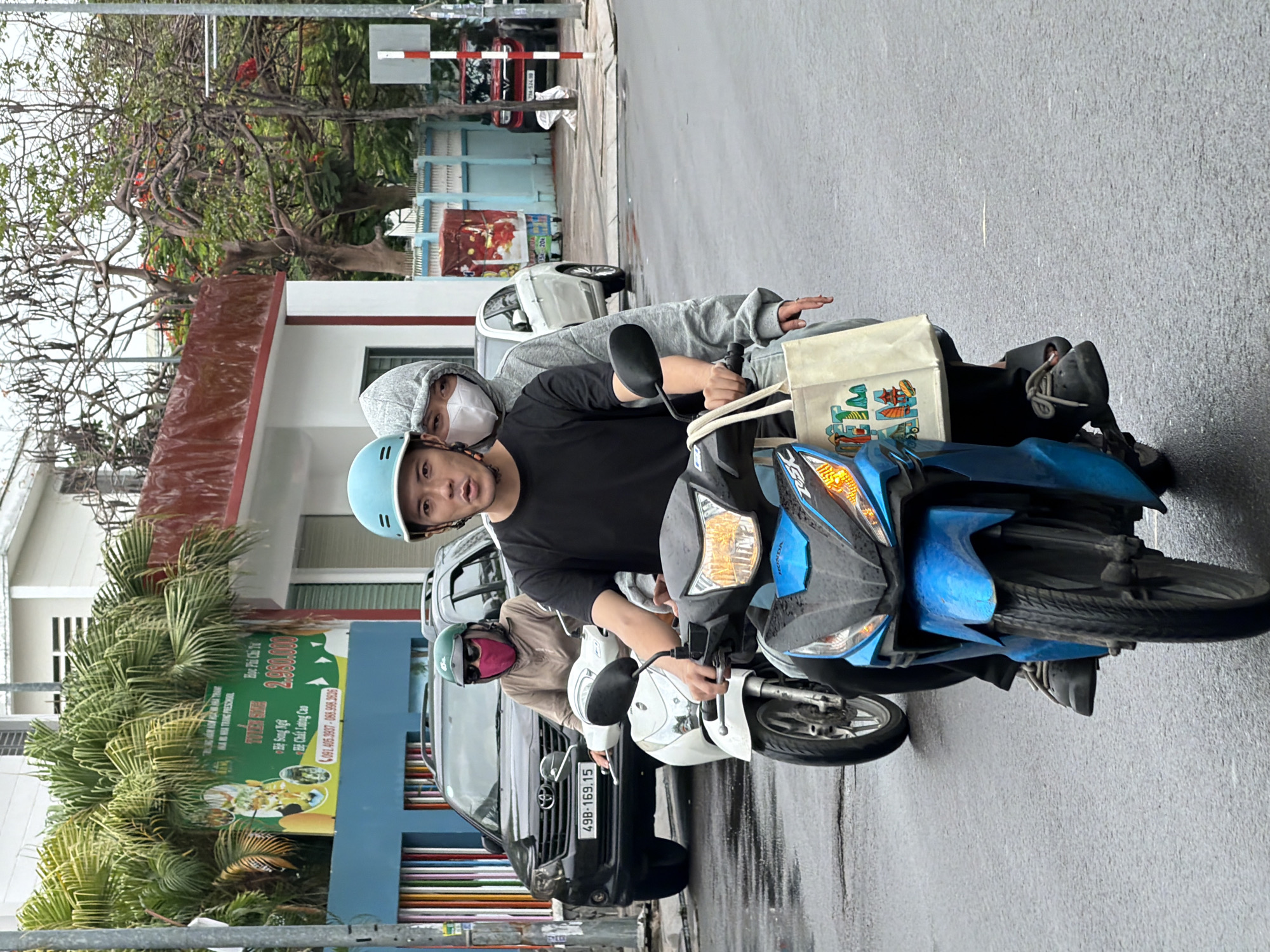 Three people are riding on two motorbikes on a street in Nha Trang, Vietnam. The leading motorbike, a blue scooter, carries two individuals. The rider, a man wearing a black t-shirt and a light blue helmet, has a concerned expression. The passenger behind him, a woman wearing a grey hoodie and a white face mask, is looking forward. A cream-colored tote bag with images of Vietnamese landmarks hangs from the handlebars of the blue scooter.
Behind them, a white motorbike is ridden by a woman wearing a green helmet, sunglasses, and a pink face covering. She is looking towards the right side of the frame.
The street is wet, suggesting recent rain. In the background, a black SUV is parked on the side of the road. Buildings with signs are visible, including one with colorful vertical stripes. Trees with green foliage and bare branches are also present, along with a street light pole. The overall scene depicts a typical urban street in Nha Trang during the daytime.