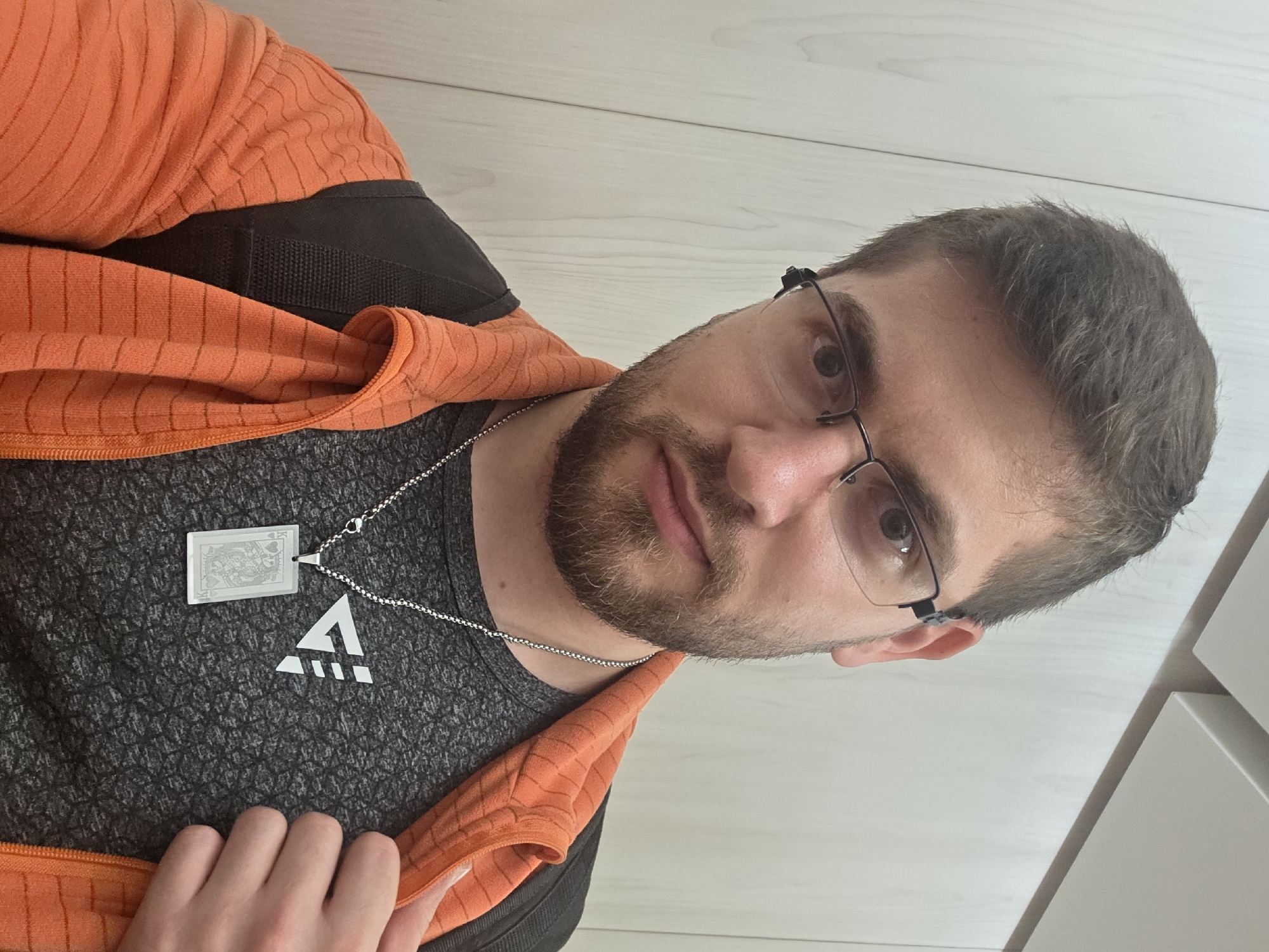 A medium-shot portrait of a Caucasian male with a short, brown beard and short, light brown hair. He is wearing glasses with thin black frames. He is wearing a gray t-shirt with a white geometric logo on the chest. Over the t-shirt, he is wearing a black vest and an orange jacket. A silver chain with two pendants hangs around his neck; one pendant appears to be a playing card and the other is a small white triangle with lines through it. He is facing the camera with a slightly downward gaze. The background is an off-white, slightly textured surface. There is no indication of the location being Plovdiv, Bulgaria.