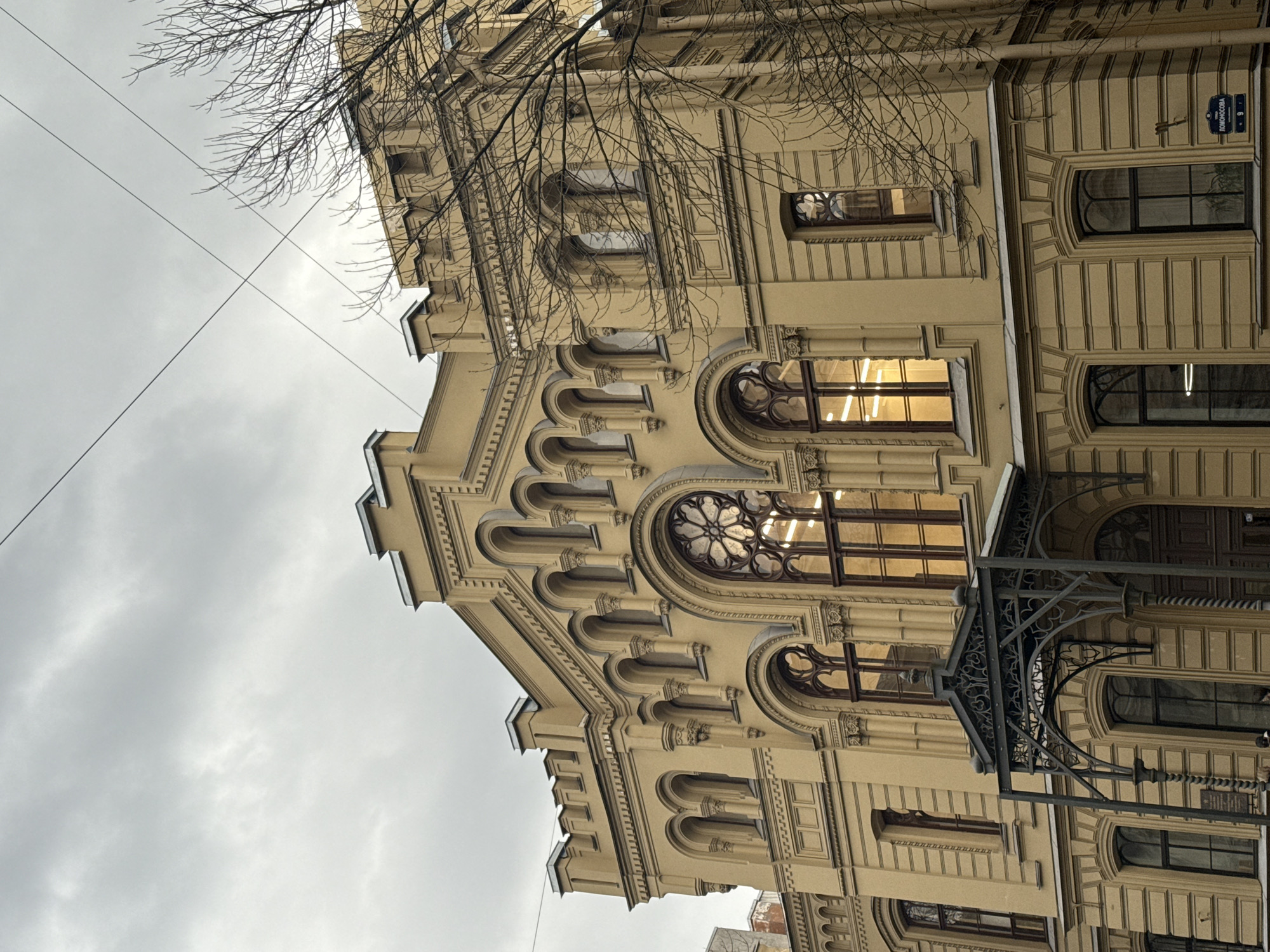 A low-angle, medium shot shows a multi-story building in Saint Petersburg, Russia, with a facade featuring elaborate, arched windows, decorative moldings, and crenelated sections. The building is painted in a beige or light yellow hue. In the upper-right corner of the frame, a street sign is visible, bearing the name "Lomonosova" and the number "9," indicating the building's address.
In the lower-right corner, a portion of a wrought-iron structure, likely a decorative canopy or entrance, is visible. A bare tree branch, devoid of leaves, extends into the upper-left portion of the frame, partially obscuring the building's upper section. The sky is overcast, with a dull grey tone. Electrical wires run diagonally across the upper portion of the scene. The windows reflect a warm, yellowish light, likely from interior illumination.