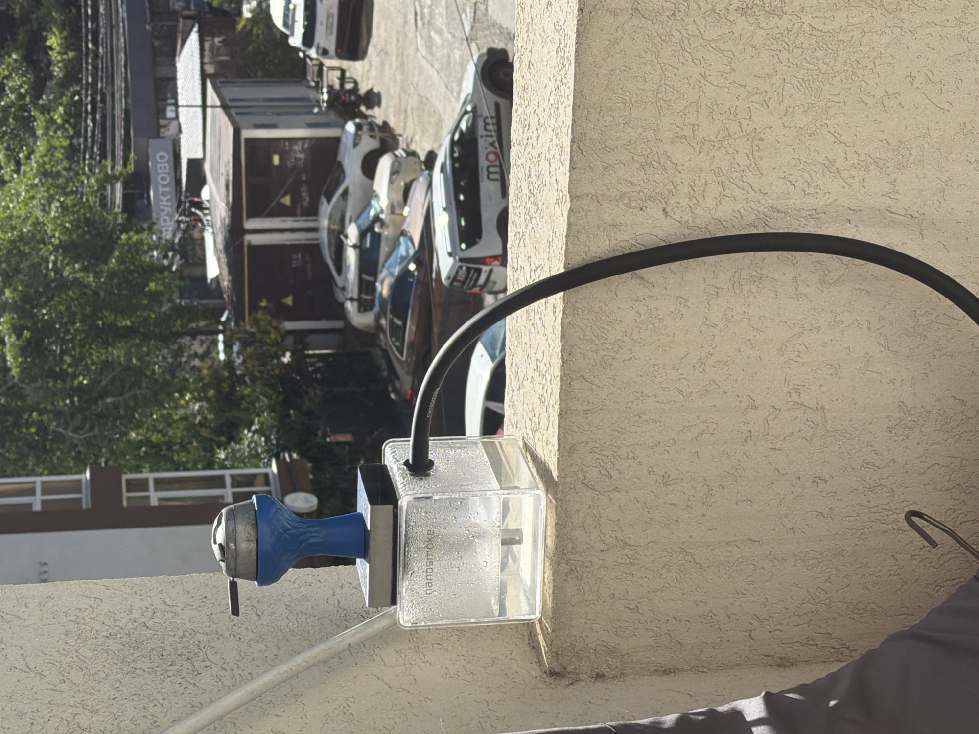 The image captures a vertically oriented outdoor scene on a bright, sunny day, featuring a mounted device in the foreground against a backdrop of a parking lot and buildings.
In the foreground, a clear, rectangular plastic box is attached to a light-colored, stucco-textured wall. The brand name "nanosmoke" is visible on the side of the box, and condensation is present inside, suggesting it contains liquid or is involved in cooling. A blue and silver metallic component is connected to the left side of this box. A thick, black flexible hose extends from the top of the box, curving upwards and to the right. Below the device, a thin, light-colored metal bar runs diagonally. A dark, possibly gray, fabric object is partially visible in the bottom right corner.
The background shows an outdoor parking area under bright daylight, indicated by strong shadows. Several cars are parked, including a prominent white car with the word "maxim" printed in black on its side, identifying it as part of the Maxim taxi service. Other white and darker-colored cars are also visible. Lush green trees line the left side of the scene. Further back, a building displays a sign with the partially visible Cyrillic text "ПРУКТОВО" (likely part of "ФРУКТОВО," meaning "fruit" or "fruit store"). The visible text "maxim" and "ПРУКТОВО" provide strong location cues, consistent with the given context of Sochi, Russia. No human activity is directly depicted.
