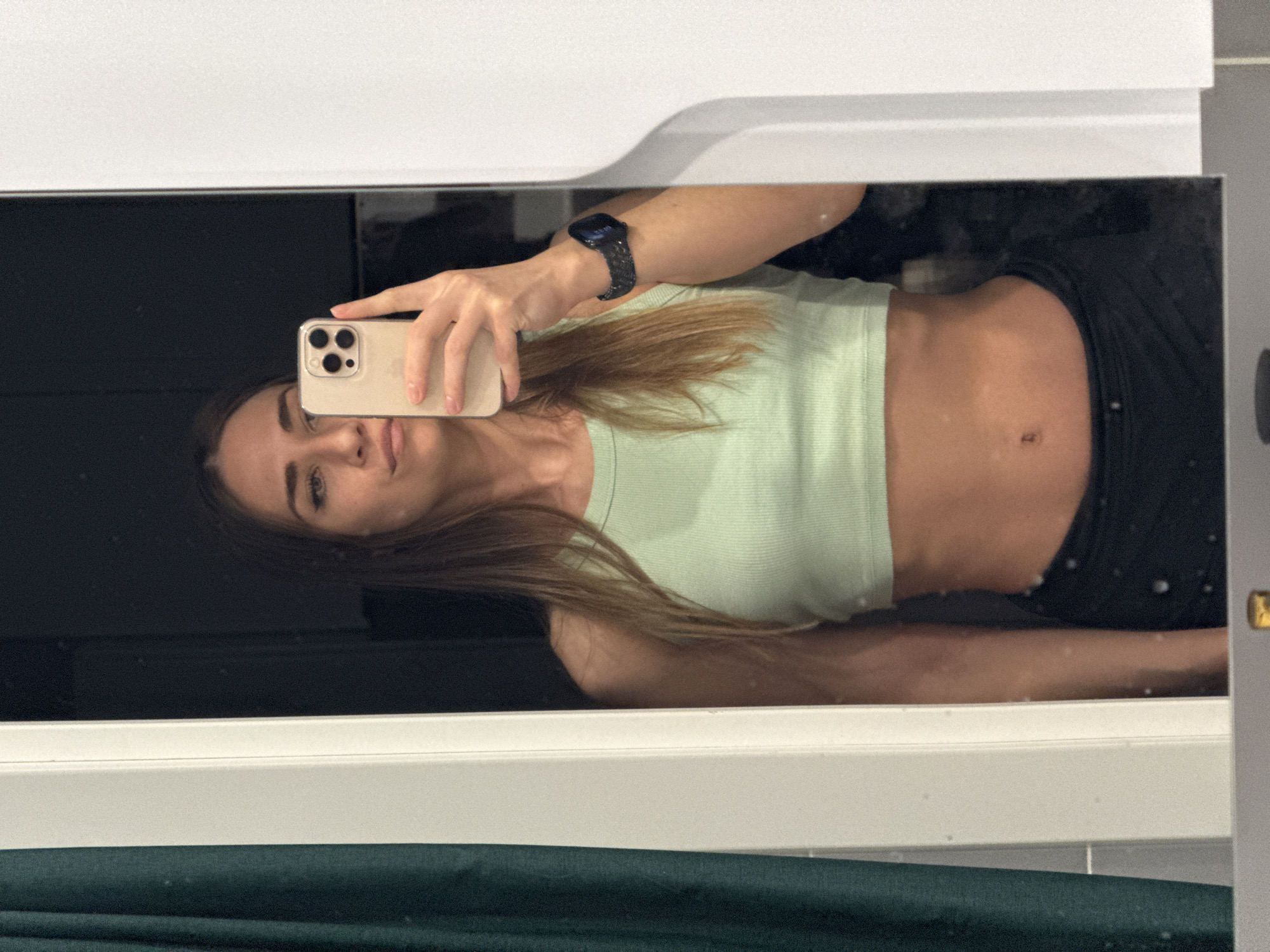 A young woman is taking a selfie in a mirror. She is wearing a light green ribbed crop top and black pants. She is holding a tan-colored smartphone in her right hand. A black smartwatch is visible on her left wrist. The mirror reflects a dark surface behind her. The mirror is framed by a white border. A dark green surface is visible below the mirror's frame. There is no information to indicate the location is Lyubertsy, Russia.