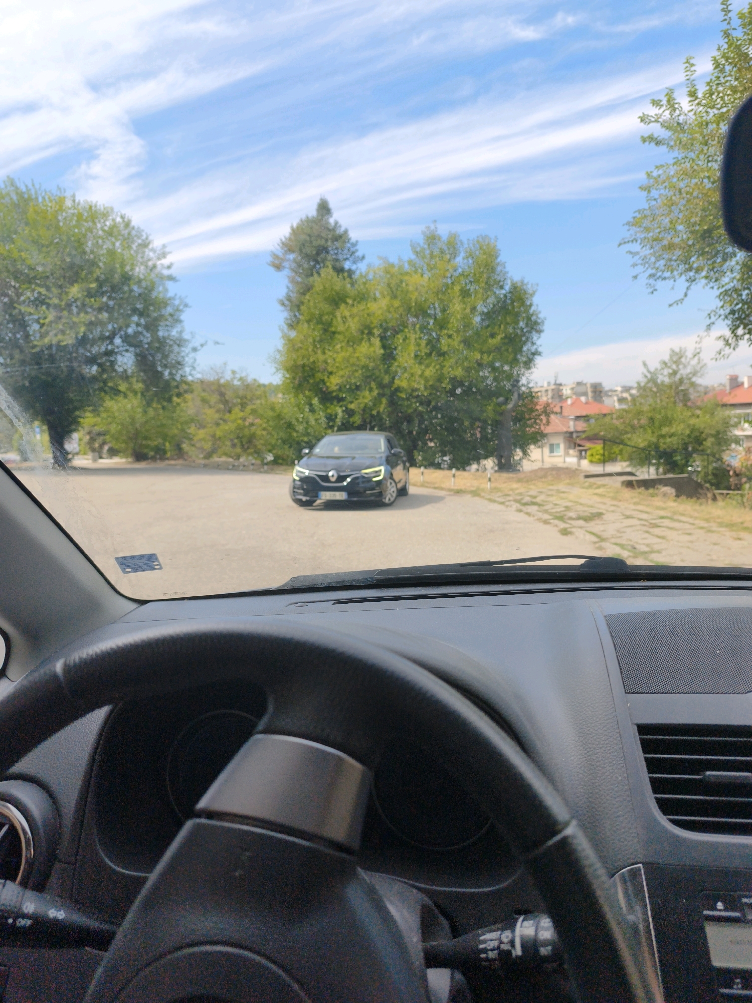 The image provides a view from inside a car, looking out through the windshield. A black sedan is parked in a paved lot in front of a line of trees and buildings. The sky is blue with scattered white clouds. The interior of the car is visible in the foreground, including the steering wheel and dashboard. The scene appears to be in a residential area, possibly in Lovech, Bulgaria, given the architectural style of the buildings.