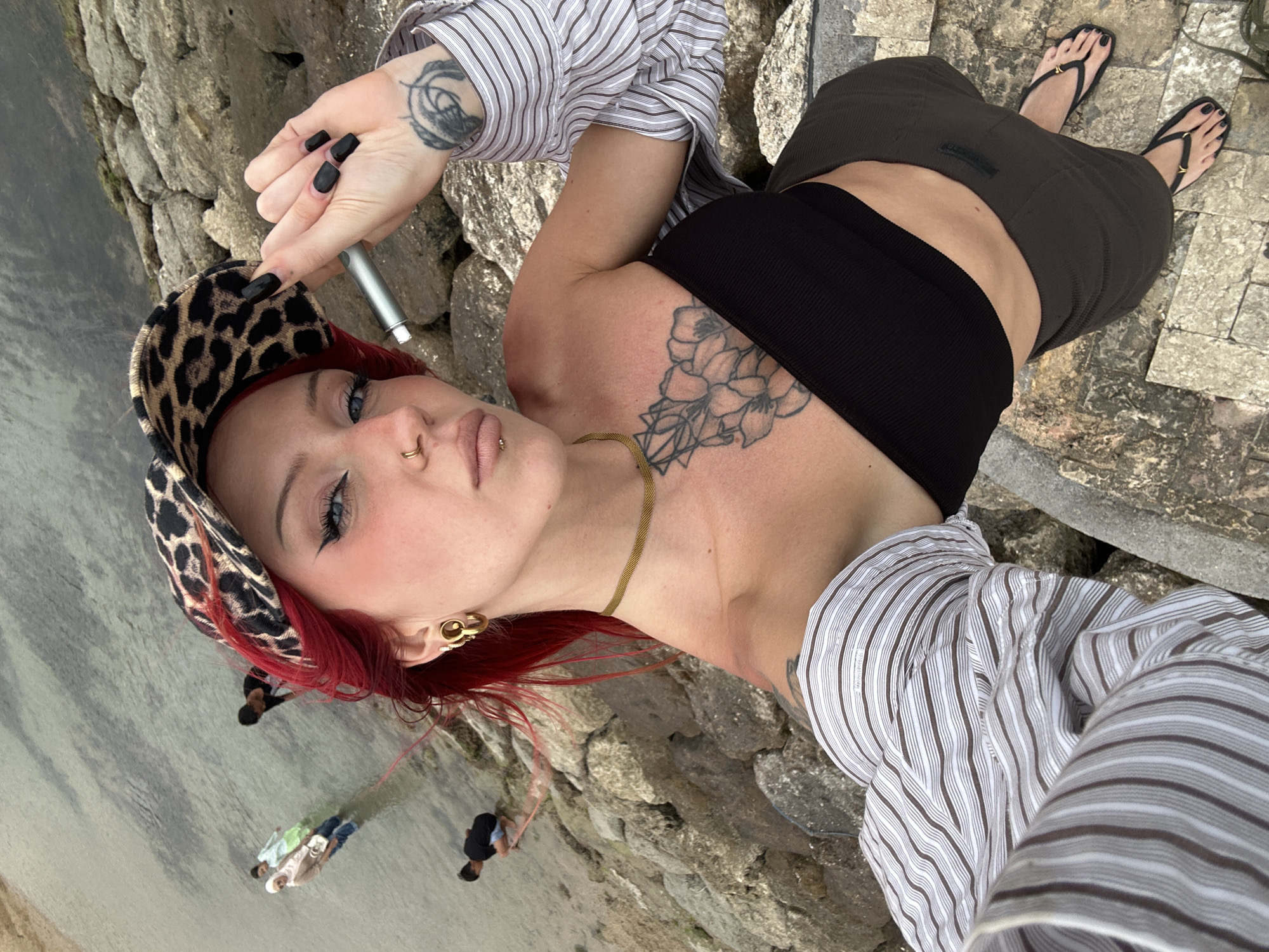 A woman with bright red hair, a leopard-print hat, and bold makeup takes a selfie outdoors. She is wearing a black bandeau top, a striped shirt unbuttoned and draped over her shoulders, and dark brown shorts. Her right hand, adorned with black nail polish and a tattoo on her wrist, holds a silver vape pen. She is also wearing large gold hoop earrings and a delicate gold necklace. Several people are visible in the shallow water in the background, including a group of three wearing headscarves. The environment appears to be a rocky shoreline or pier, with the ocean or a large body of water to the left and stone steps leading down to the right. The rocky texture and the presence of the water suggest a coastal location. The specific location in Denpasar, Indonesia, is not discernible from the image.
