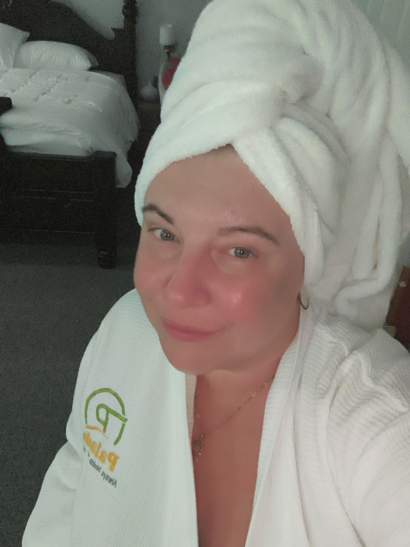 A woman with a white towel wrapped around her head and a white robe is taking a selfie in a room. She has light skin and is wearing a gold necklace. The robe has a green and yellow logo on the left side. In the background, there is a bed with white sheets and a dark wooden headboard, and a nightstand with a lamp. The room appears to be dimly lit. The location is not identifiable as Phu Thuy Ward, Vietnam.