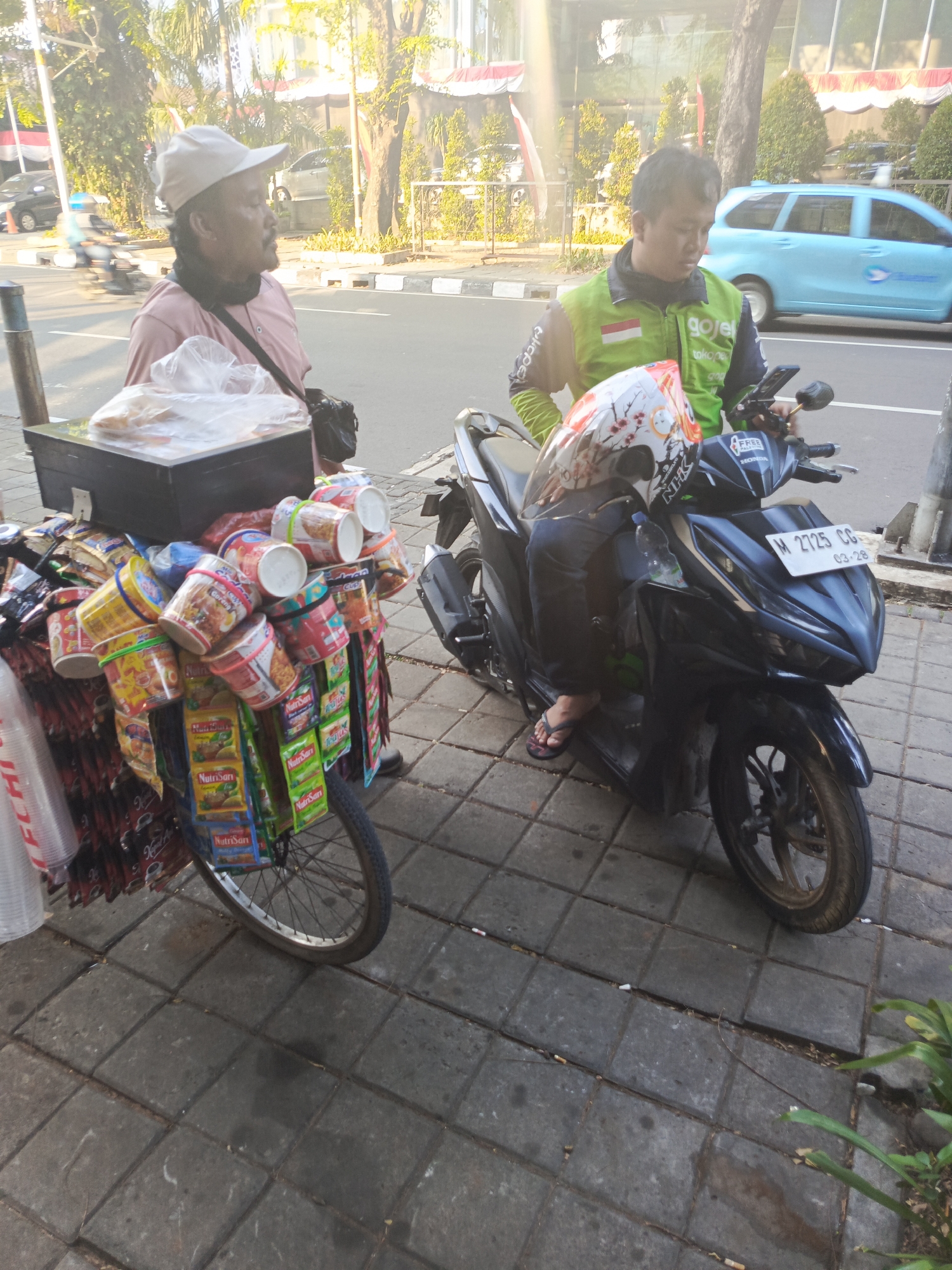 A man in a pink shirt and a cap stands next to a mobile food cart laden with packaged snacks and drinks. He appears to be engaged in conversation or transaction with a man wearing a green Gojek jacket who is seated on a black motorcycle. The motorcycle has a license plate reading "M 2725 CG" and "03-28". In the background, a light blue car is visible on the road, along with trees and buildings. The scene takes place on a sidewalk made of interlocking gray paving stones. The overall lighting suggests it is daytime, possibly morning or late afternoon.
