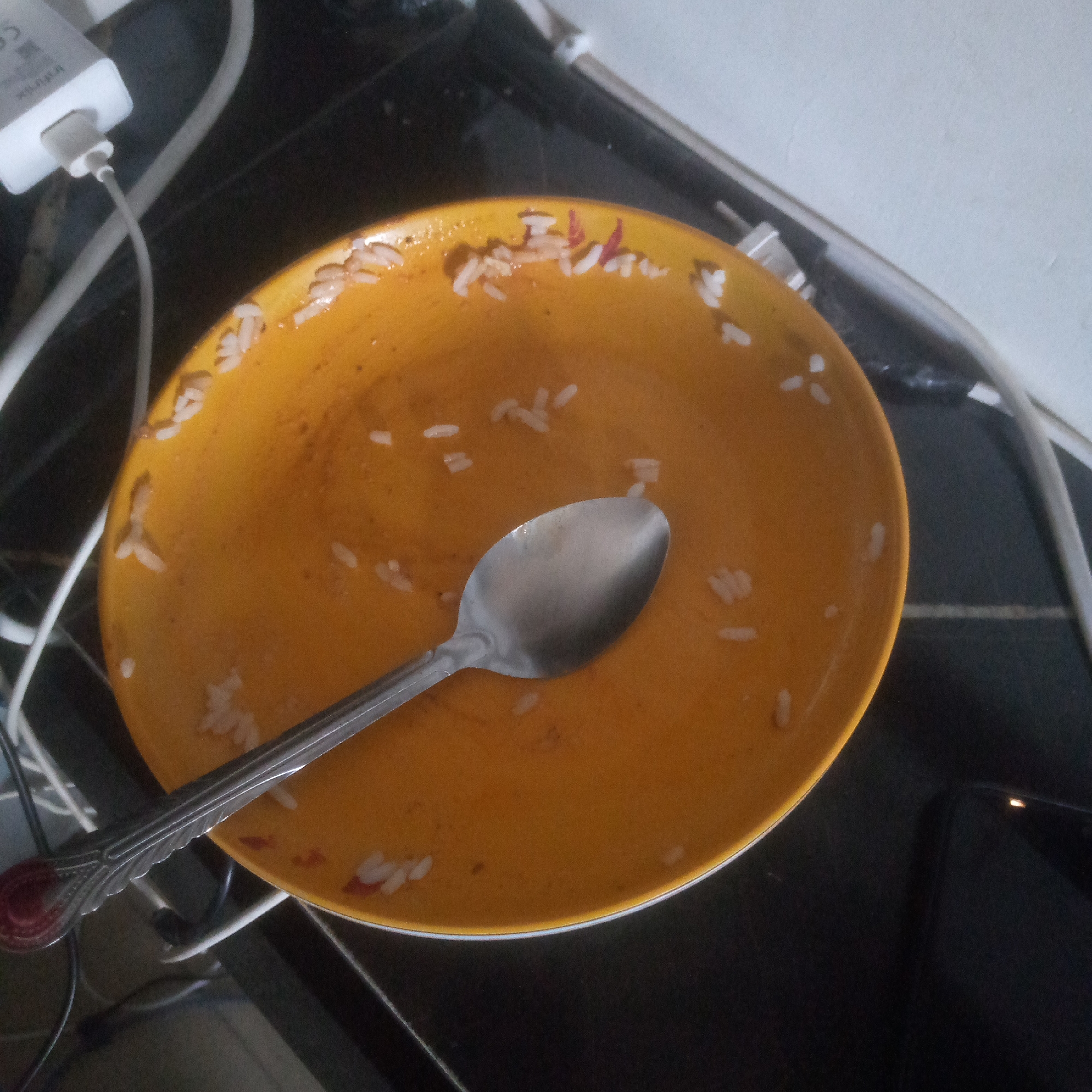 An overhead, slightly angled medium shot captures a dirty, yellow, circular plate with remnants of food and a spoon on a dark, reflective surface. The plate, positioned in the center, contains scattered white grains of rice and a few red flecks, possibly food debris. A silver spoon rests on the plate, with its bowl positioned in the upper right quadrant of the plate and its handle extending towards the bottom left. In the background, to the left of the plate, a white electronic device with a USB port is visible, connected to a grey cable. The surface beneath the plate appears to be a dark, possibly black, countertop or table. The lighting is dim, with some glare reflecting off the surfaces. The overall impression is of a used plate after a meal, with the setting suggesting an indoor, domestic environment.