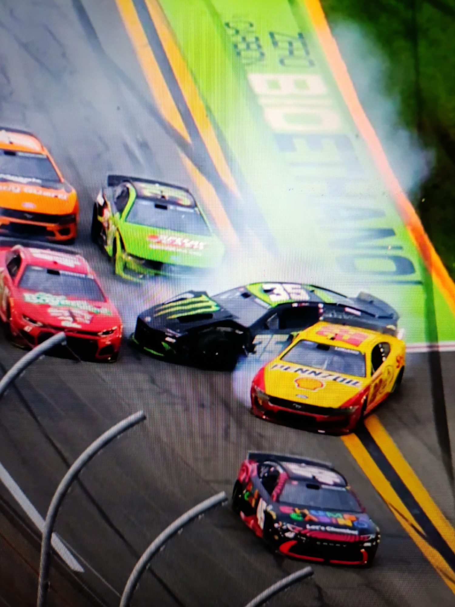 In an aerial view of a race track, several stock cars are depicted in motion. One car, colored orange, is in the foreground, followed by a green and white car. To the left and slightly behind these, a red car is visible. A black car with a green monster logo is positioned behind the red car, and a yellow and red car is to its right. A black car with colorful decals is at the bottom right of the frame. The race track itself is marked with yellow lines, and a green wall with white lettering is visible in the background. The image appears to be a capture of a racing event.