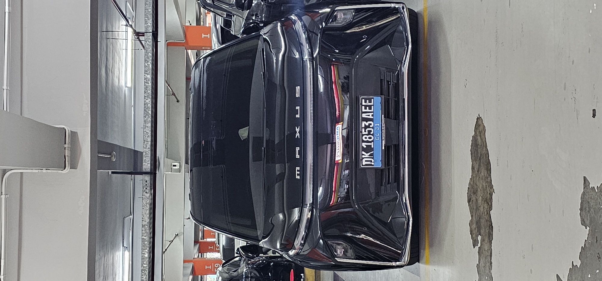 A vertically-oriented photograph depicts the rear of a dark-colored Lexus vehicle parked in what appears to be an indoor parking garage. The vehicle’s license plate, "DK 1853 AEE", suggests that the location is in Bali, Indonesia, where "DK" is a common license plate prefix. The expiration date on the license plate is "04 21". The Lexus emblem is visible on the rear of the car, spelled out vertically. Overhead lighting fixtures and structural supports of the parking garage are visible in the background. The concrete floor shows signs of wear and tear. There are parking spot markers, some colored orange, visible in the background. The presence of the Lexus and the parking garage environment indicate a modern setting.
