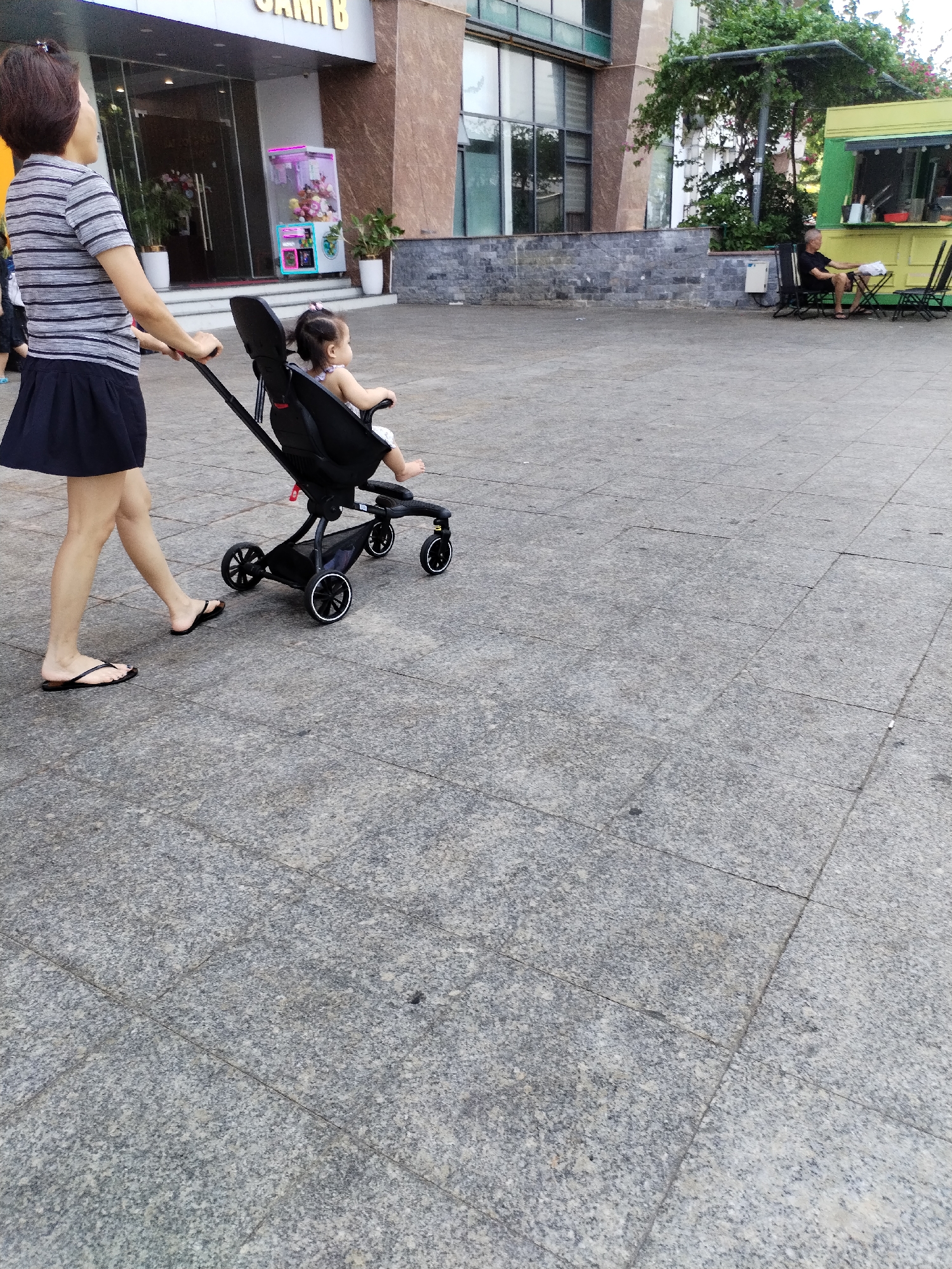A woman in a striped shirt and black skirt walks to the right, pushing a black stroller with a baby inside. The baby is wearing a white outfit and has a pink bow in its hair. They are on a paved area with square grey tiles. In the background, a building with large windows and a sign reading "CANH B" is visible. A green food truck is parked to the right, and a man in a black shirt and shorts is sitting on a chair nearby. The setting appears to be an urban outdoor space, possibly a plaza or park area in Nha Trang Ward, Vietnam.