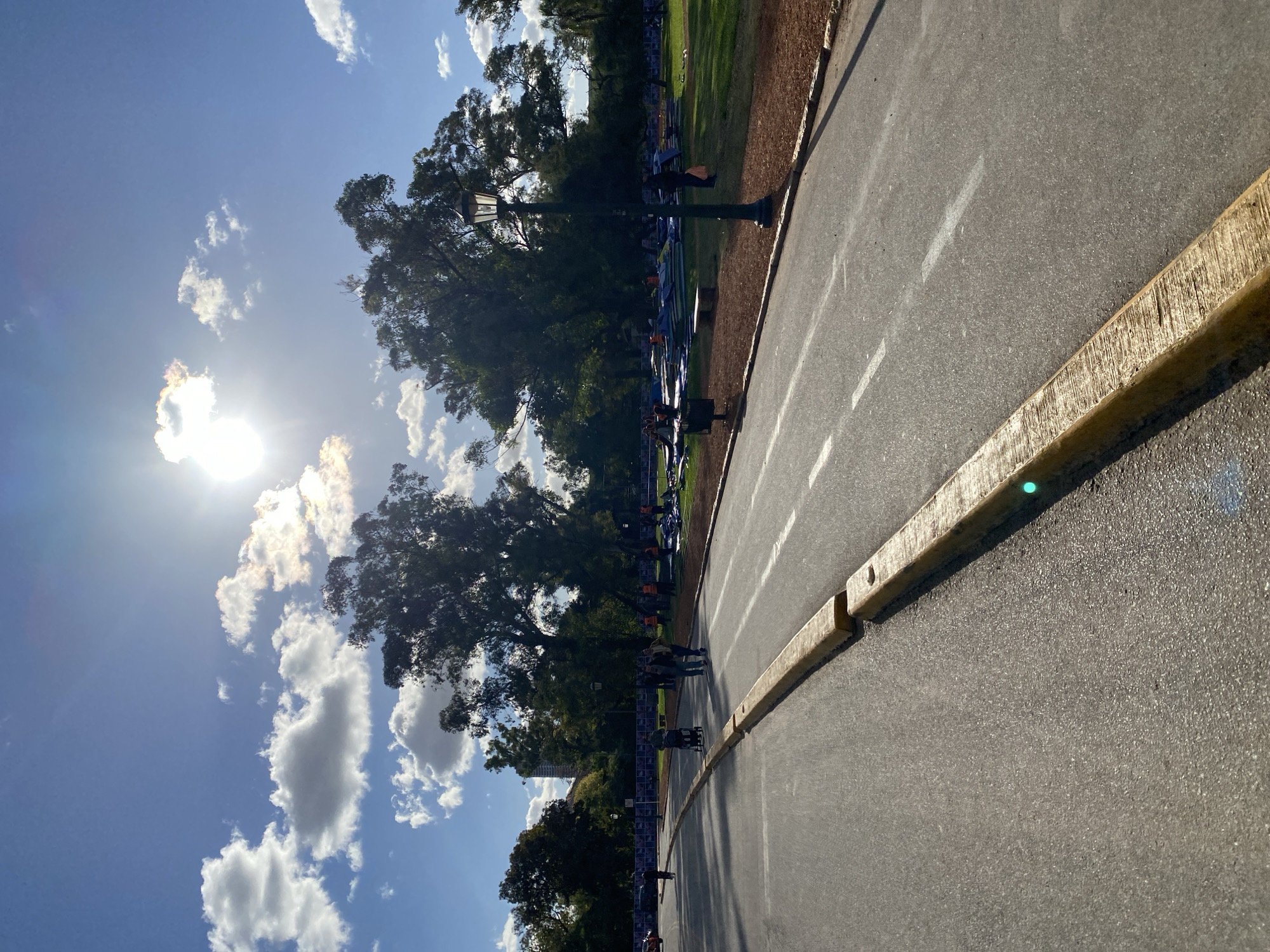 This image captures an outdoor scene, likely a park or a wide avenue, under a bright blue sky in Buenos Aires, Argentina. The foreground features a paved road or track with white dashed lines, bordered by a textured yellow concrete barrier or curb.
To the left of the road, there's a strip of brown earth or mulch, followed by a grassy lawn shaded by several large, leafy trees. Along the edge of the grassy area, temporary blue and white event barriers or fencing are set up, behind which various blue and white inflatable structures or tents are visible. These elements suggest an organized outdoor event is taking place or has recently occurred.
Several people are scattered throughout the scene, walking or standing, predominantly near the event barriers and under the trees. One person is visible near a tall, dark green lamppost with an ornate top, which stands between the grassy area and the road.
The time of day appears to be mid-morning or early afternoon, characterized by intense sunlight high in the sky, causing a strong sunburst effect and lens flare on the left side of the image. The weather is clear and sunny, with scattered white cumulus clouds across the blue sky. The overall atmosphere is bright and calm, implying a pleasant day for an outdoor gathering. No visible text is discernible on any objects or structures.
