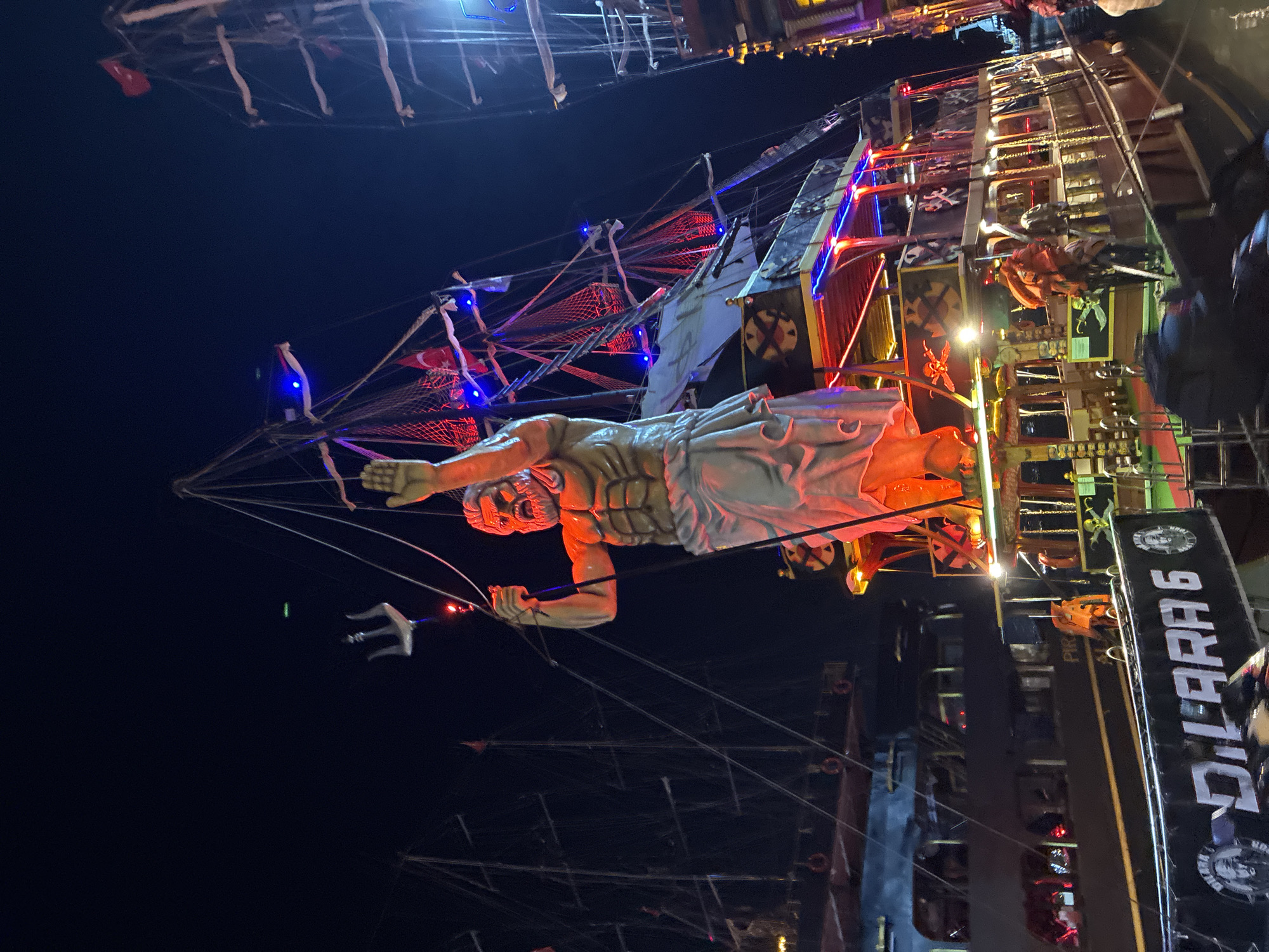 The image depicts a nighttime scene in Alanya, Turkey, featuring several elaborately decorated pirate-themed boats docked together. The most prominent element is a large, illuminated statue of a muscular, bearded male figure, resembling a mythological deity or a powerful pirate captain, standing atop one of the boats. This statue is lit with red and orange lights, highlighting its physique and a flowing white garment. The figure holds a trident in its raised left hand.
The boat the statue is on is adorned with pirate motifs, including skull and crossbones flags and crossed swords. It is further decorated with numerous colorful LED lights, predominantly red and blue, illuminating the railings, decks, and sails. The boat itself has multiple levels with railings and what appear to be seating areas. Several people, some wearing orange life vests, are visible on the deck, suggesting a potential tour or event.
Behind the main boat, other pirate-themed vessels are visible. These also feature tall masts, rigging, and flags, some of which display the Turkish flag. These boats are also illuminated, creating a festive and lively atmosphere against the dark night sky. The water surface is visible at the bottom of the frame, reflecting some of the lights. The overall impression is one of a vibrant entertainment venue or a tourist attraction in Alanya. The name "DİLARA 6" is visible in large white lettering on the side of one of the boats, potentially indicating its name or identifier.
