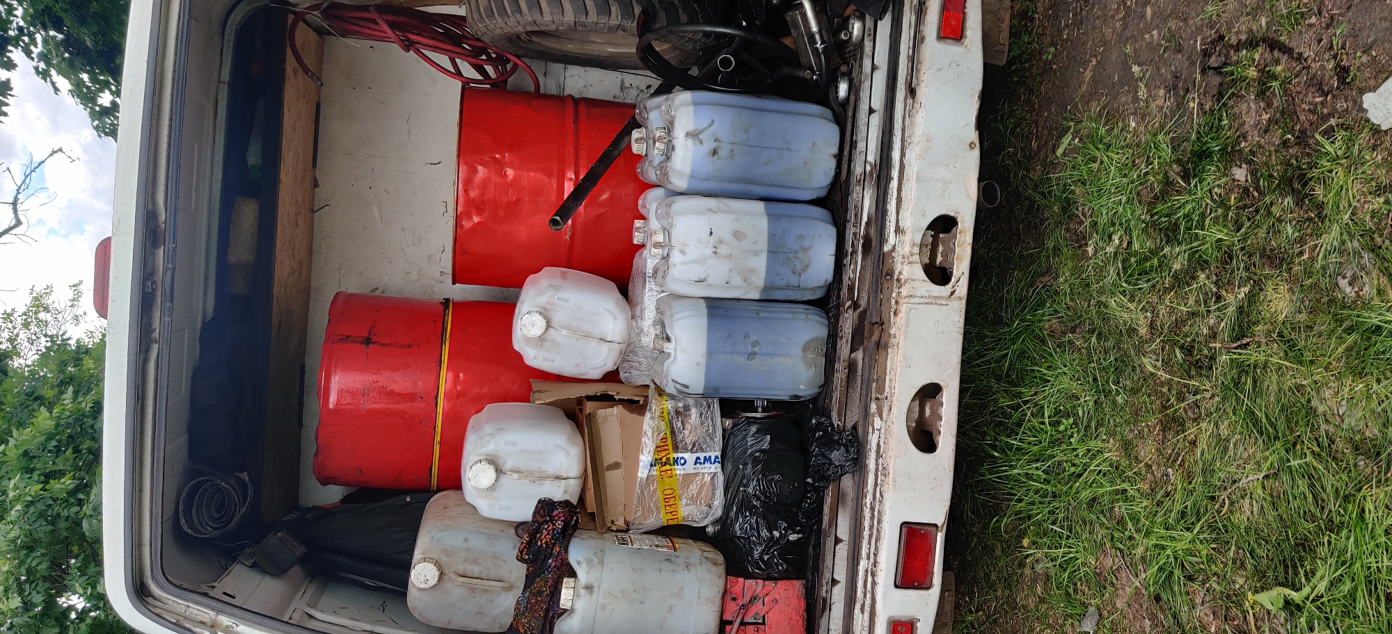 The image shows the open rear of a white van or truck, heavily laden with various items. The interior appears to be filled with cargo, including two red metal barrels, multiple white plastic jerrycans (some with a blue liquid), a spare tire, a steering wheel, a black plastic bag, and cardboard boxes wrapped with tape. The vehicle is parked on grass. The location is in an undefined rural area. There are no people visible in the image.