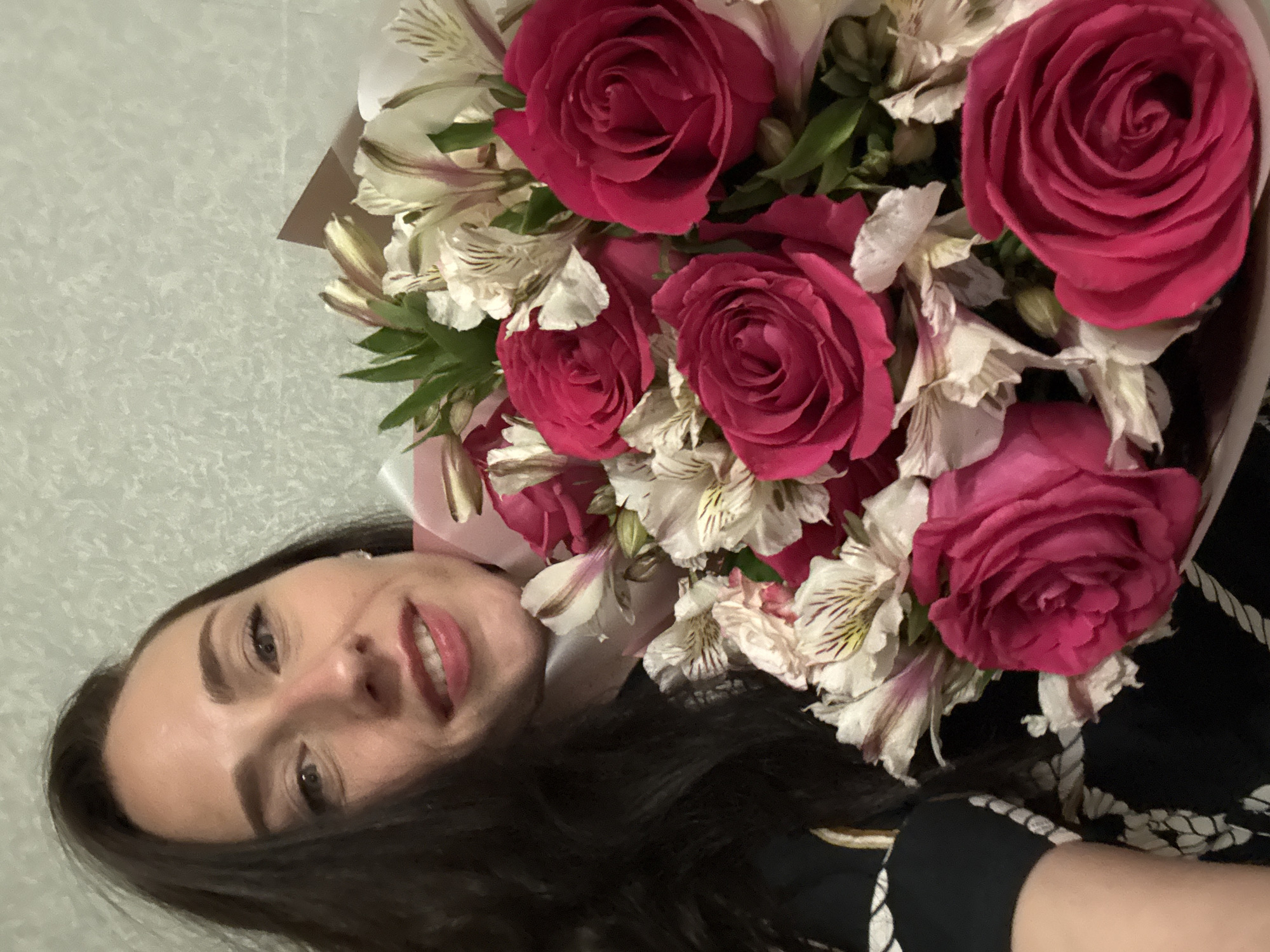 A woman is holding a bouquet of flowers. The bouquet contains pink roses and white alstroemeria lilies. The woman has dark hair and is smiling. The background is a light-colored wall. There are no other people or objects visible in the image. The location is unknown.