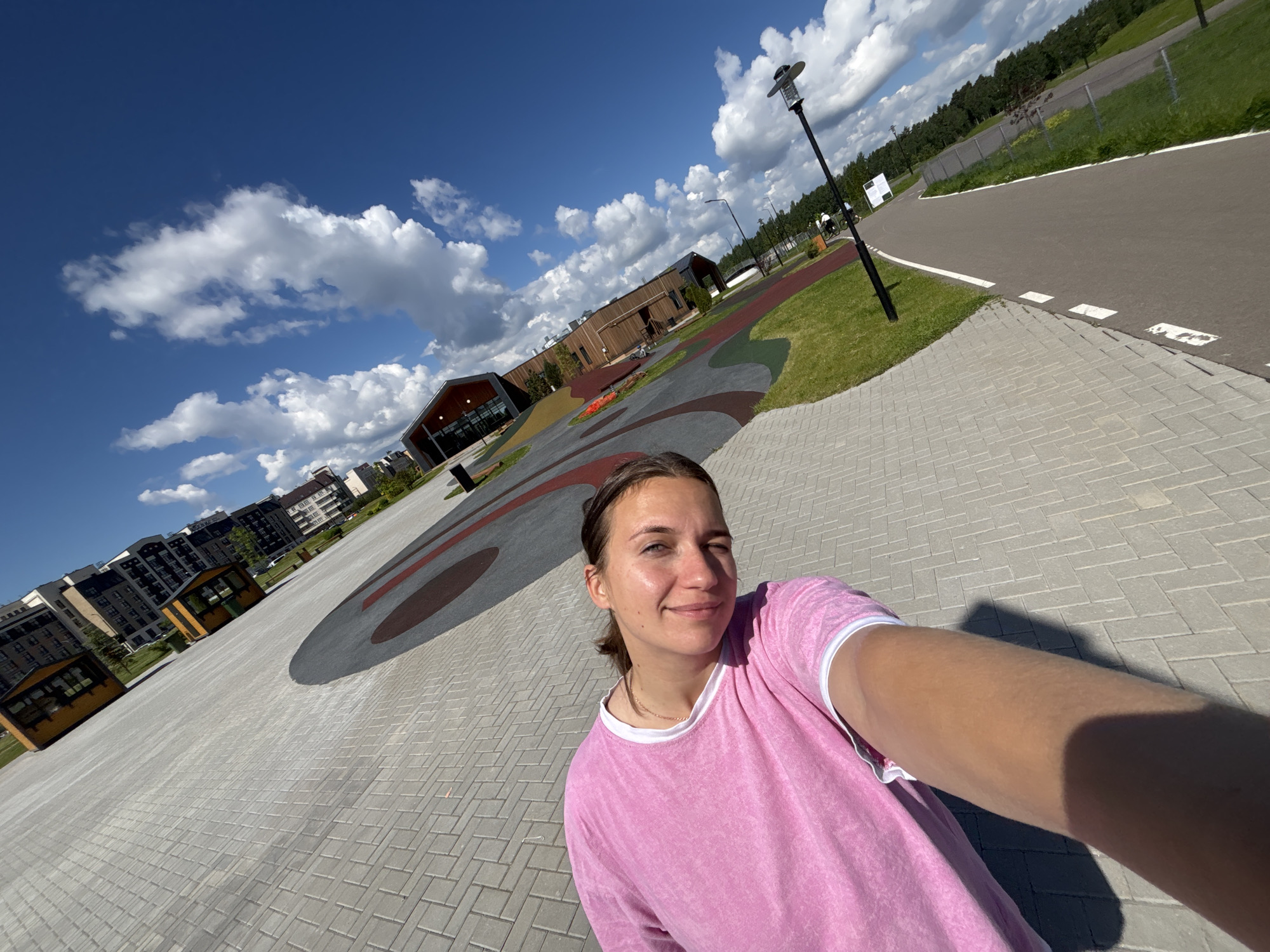 A selfie taken in Varaksino, Russia, shows a young woman with brown hair pulled back, wearing a pink t-shirt with white trim, smiling slightly at the camera. The background consists of a pedestrian area paved with grey interlocking bricks, a playground area with a large circular design on the ground in shades of grey, brown and red, modern low-rise residential buildings, and a blue sky with scattered white clouds. A bicycle path and a lamppost are also visible on the right side of the image. The lighting indicates it is daytime.