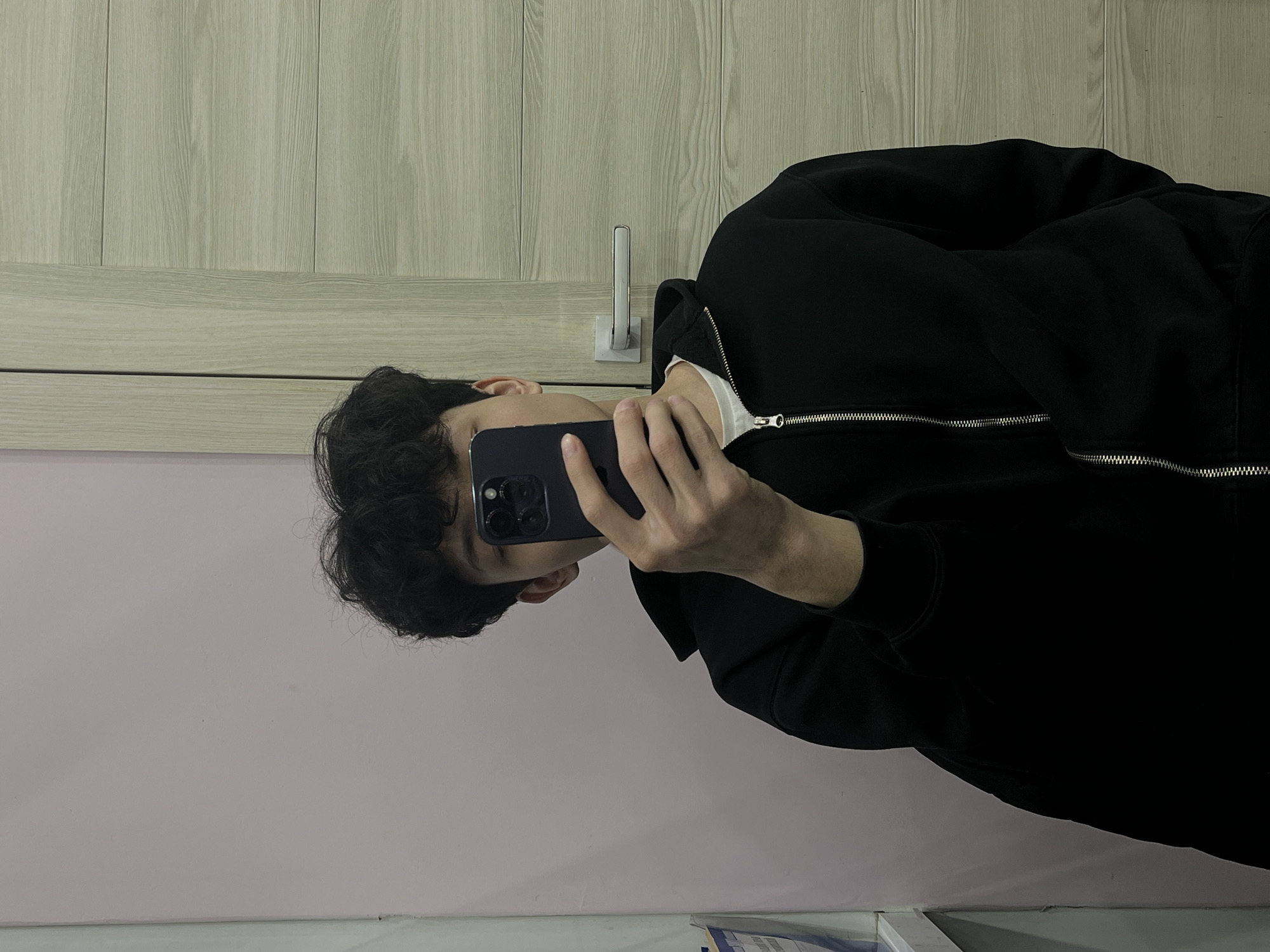 A person with curly dark hair is taking a selfie with a dark smartphone, partially obscuring their face. The person is wearing a black zip-up hoodie over a white undershirt. The background consists of a pink wall, a vertically paneled wall, and what appears to be a mirror reflecting the scene. A wall-mounted accessory is also visible on the paneled wall.