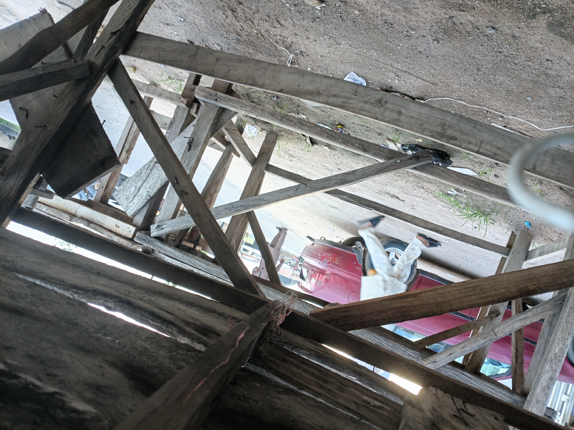 The image depicts an outdoor scene in Jalingo, Nigeria, characterized by a makeshift wooden structure in the foreground. The structure appears to be under construction or in disrepair, with numerous rough-hewn wooden planks forming a canopy or shelter. Sunlight filters through gaps in the wooden beams, creating streaks of light and shadow on the ground.
In the mid-ground, a red car is partially visible, suggesting it is parked or passing by. A person wearing light-colored clothing, possibly traditional attire, is walking towards the right side of the frame. Their legs and feet are visible, wearing dark sandals. Further in the background, another individual in similar attire can be seen walking away from the camera. The ground is covered in dirt and gravel, with sparse patches of dry grass and some scattered debris, including plastic bags and wrappers. The overall environment suggests a rural or semi-urban setting within Jalingo. The perspective is from low down, looking up into the wooden structure.