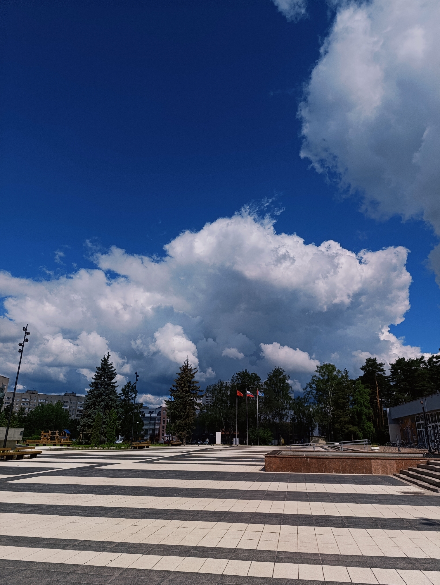 This media file depicts an outdoor scene in Chernogolovka, Russia. The foreground features a spacious plaza paved with alternating black and white striped tiles, creating a striking geometric pattern. Several dark wooden benches with metal frames are placed along the edges of the plaza.
In the middle ground, there are evergreen trees, including tall fir trees, and deciduous trees. A modern building with a gray facade and large windows is visible on the right side. To the left of the trees, a multi-story residential building with a light-colored facade and numerous windows can be seen. Three flags are prominently displayed on flagpoles: one appears to be the Russian flag, another is red, and the third is blue with some emblem.
A low-rise building with a reddish-brown granite base, possibly a fountain or water feature, is situated in the middle of the plaza. A lamppost with multiple lights stands to the left of the trees. The sky is a vibrant blue, filled with dramatic, large white cumulus clouds, suggesting it is daytime, likely mid-afternoon, with fair weather.
There are no people clearly visible in the frame, though a hint of activity might be suggested by bicycles parked near the building on the right. The overall mood is serene and open, highlighting the architectural design of the plaza and the natural elements of the trees and sky. The location is identified as Chernogolovka, Russia.